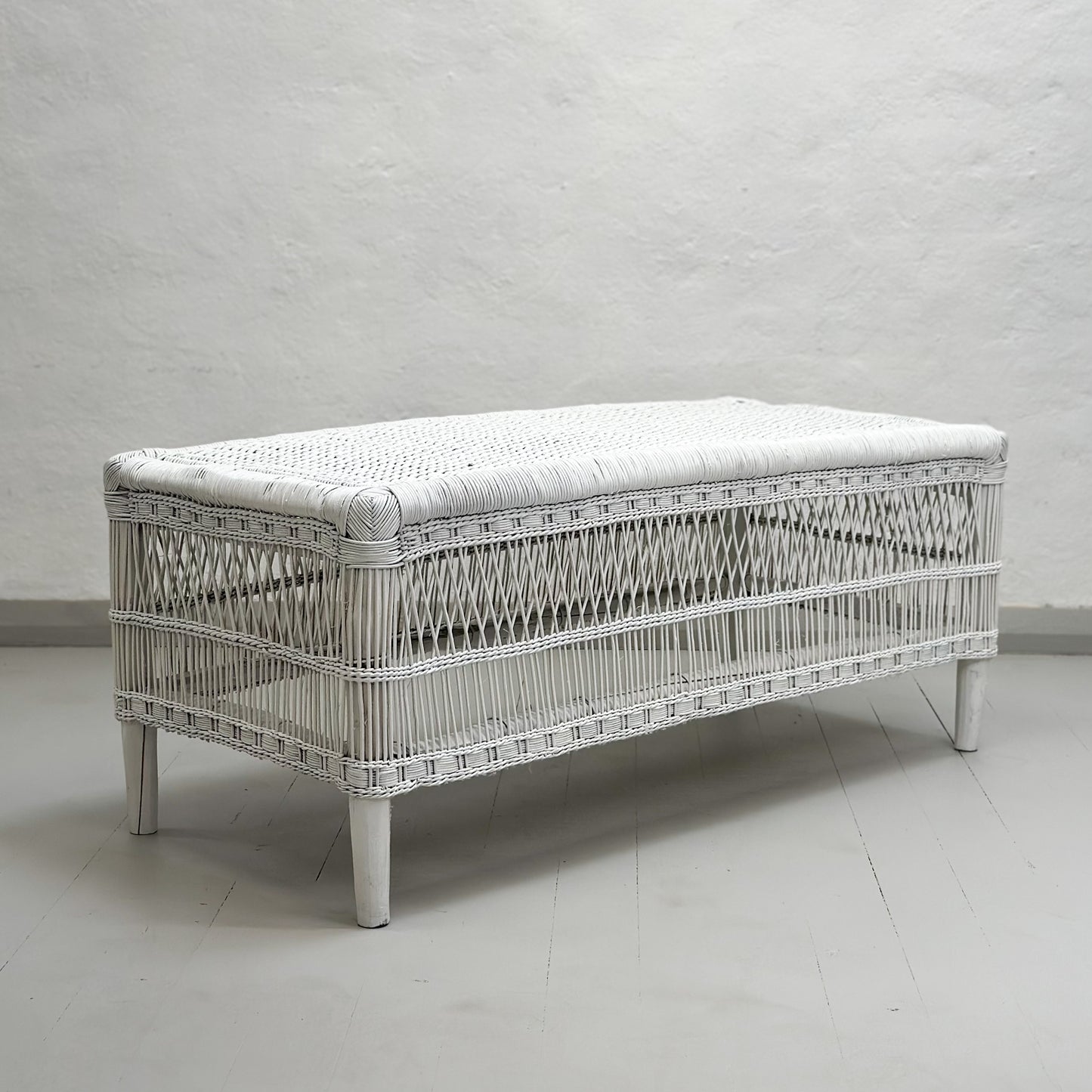 A handwoven white Malawi cane bench with a flat base, depicted in a room with light grey walls.