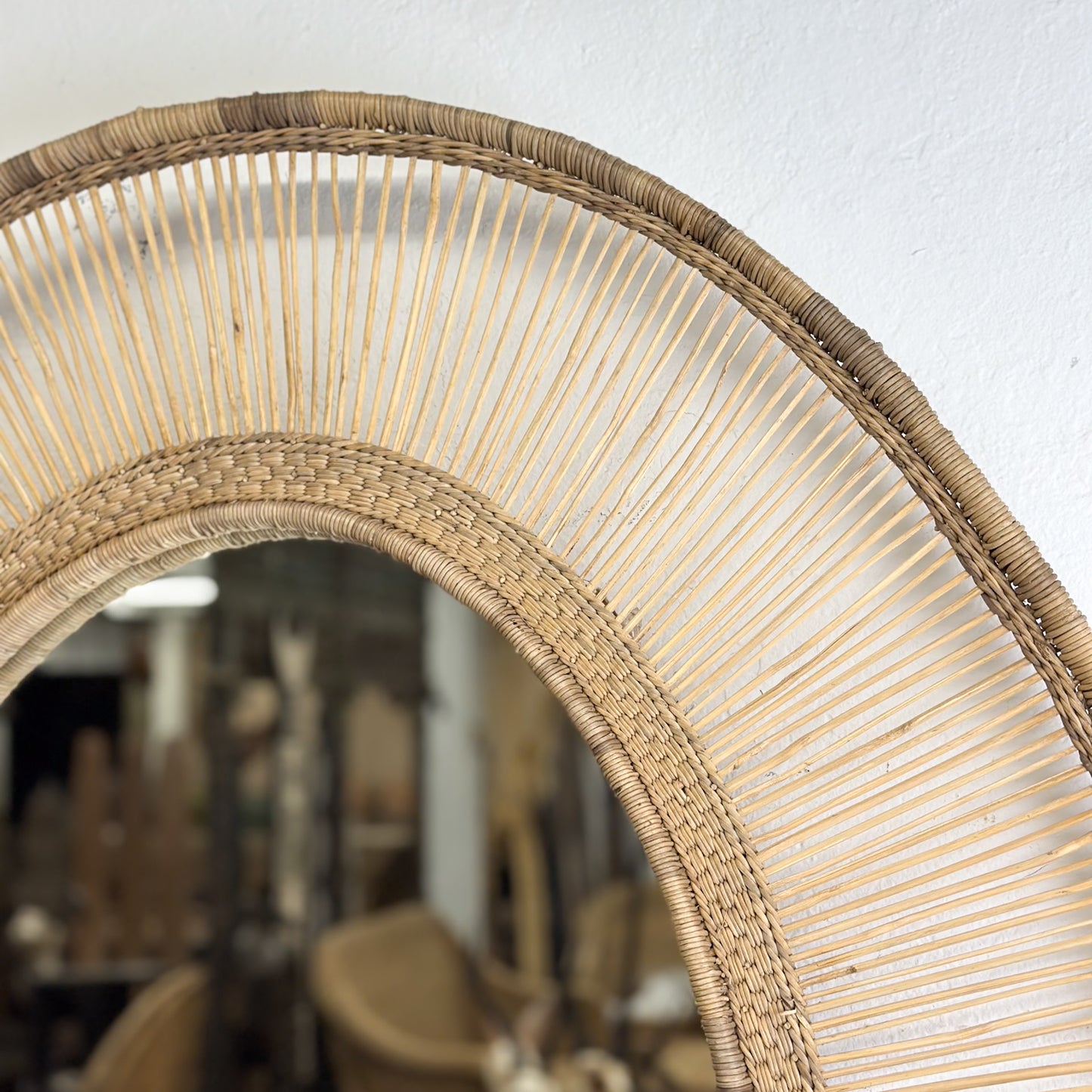 An oval-shaped mirror with a natural, woven frame, reflecting a room with white walls and a wooden chair.