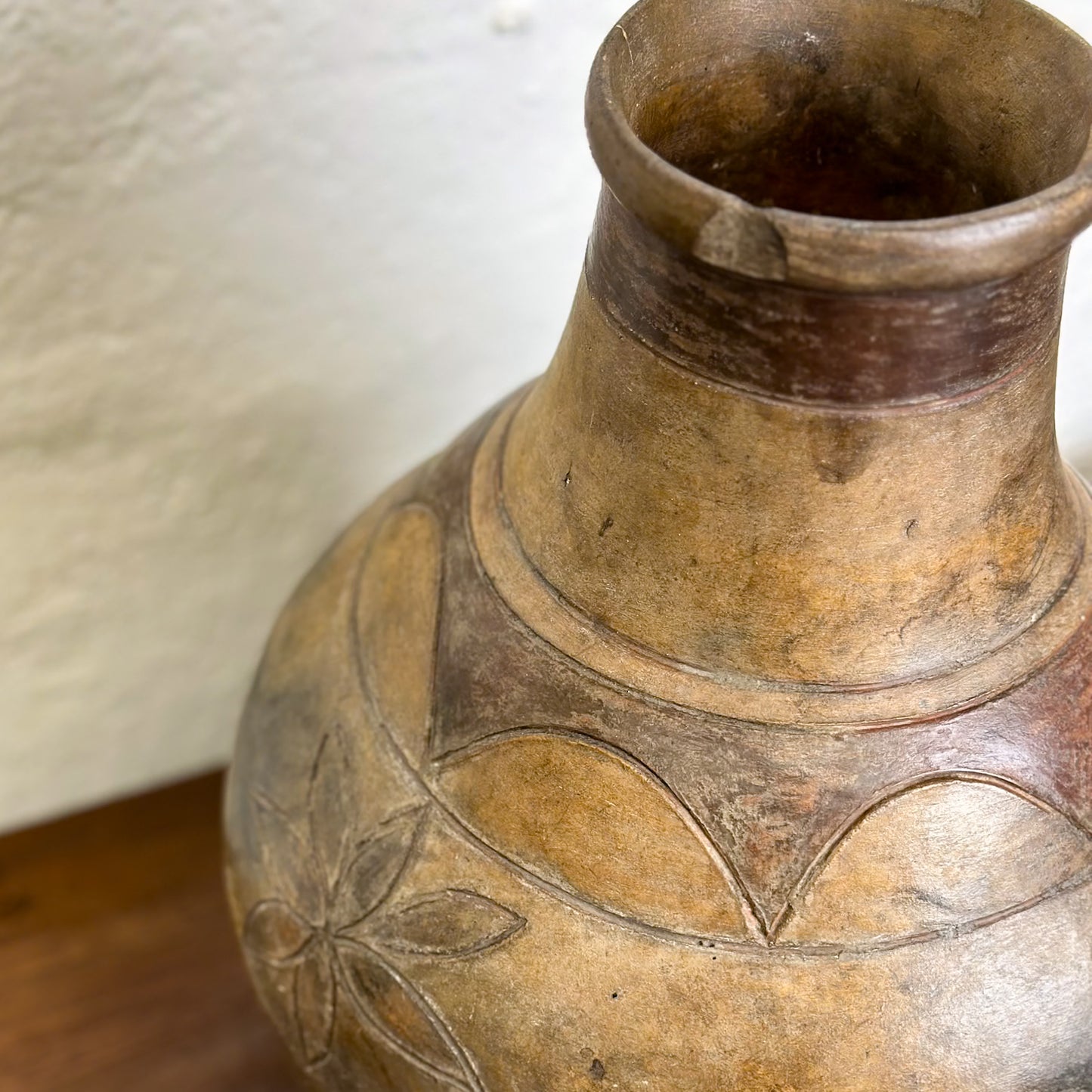 A brown ceramic pot with incised geometric patterns and a lid, placed on a wooden surface against a white wall.