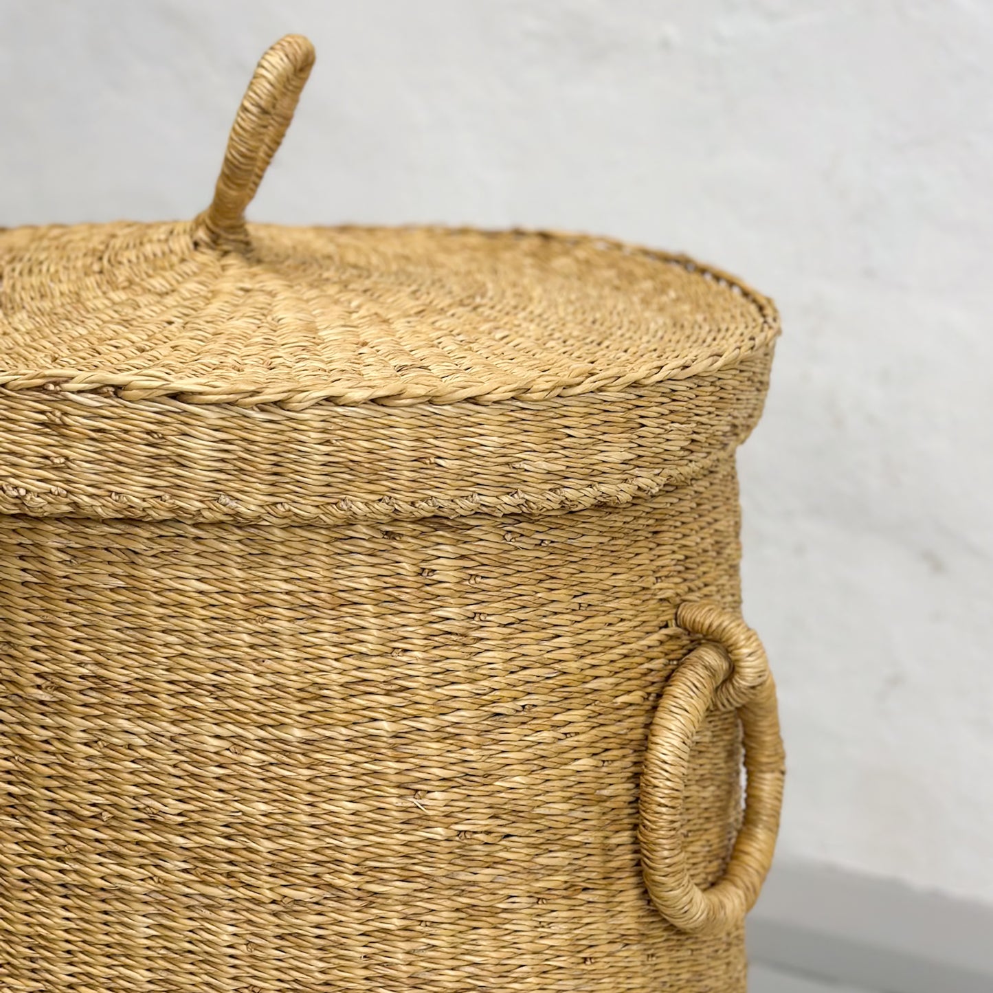 A handcrafted laundry basket made of wicker, with a natural beige color, featuring two handles and a lid, set against a plain background.