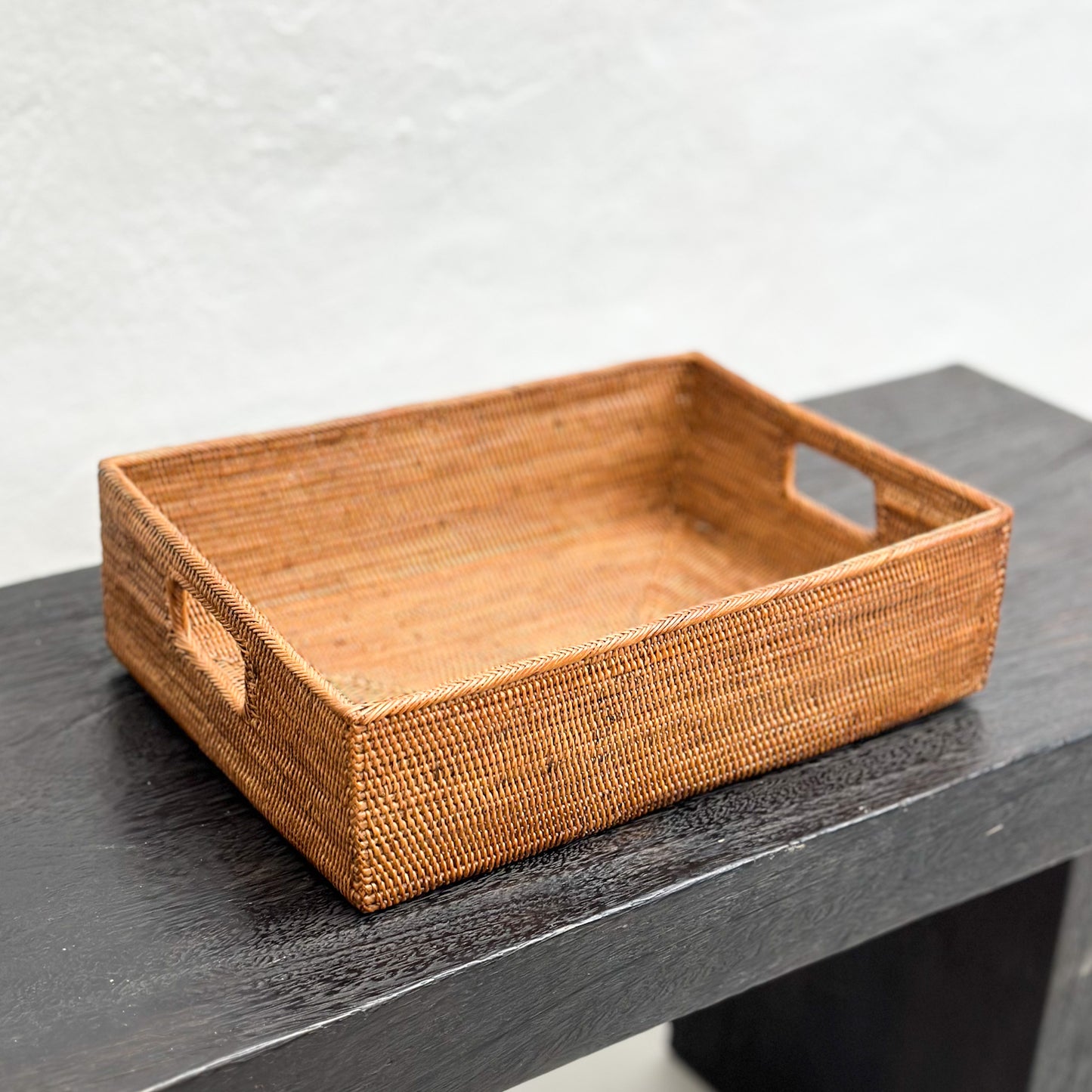A rectangular wicker towel tray basket with handles, placed on a dark surface with a plain background.