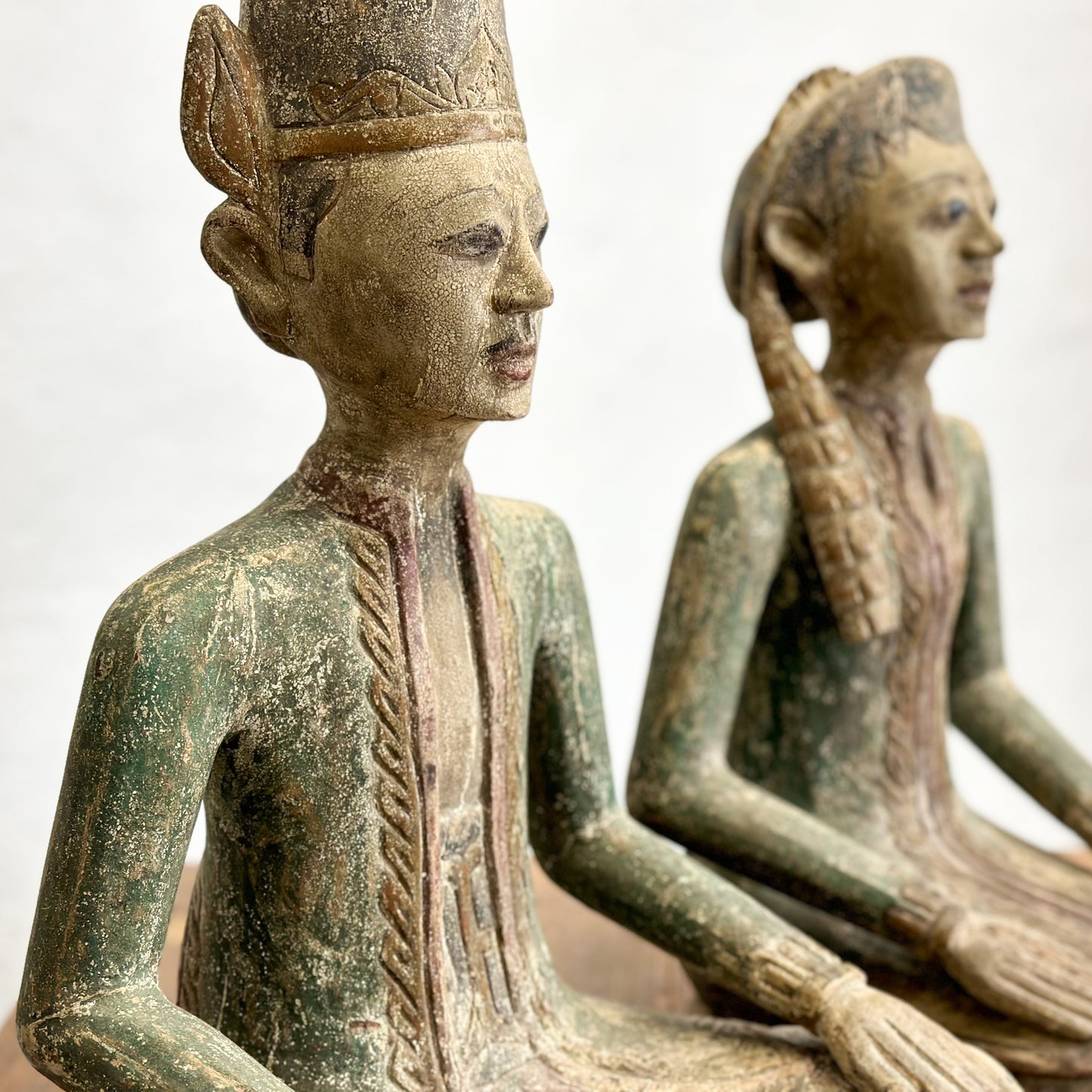 Two vintage carved wooden statues of seated figures with decorative heads, placed on a wooden surface against a white wall.