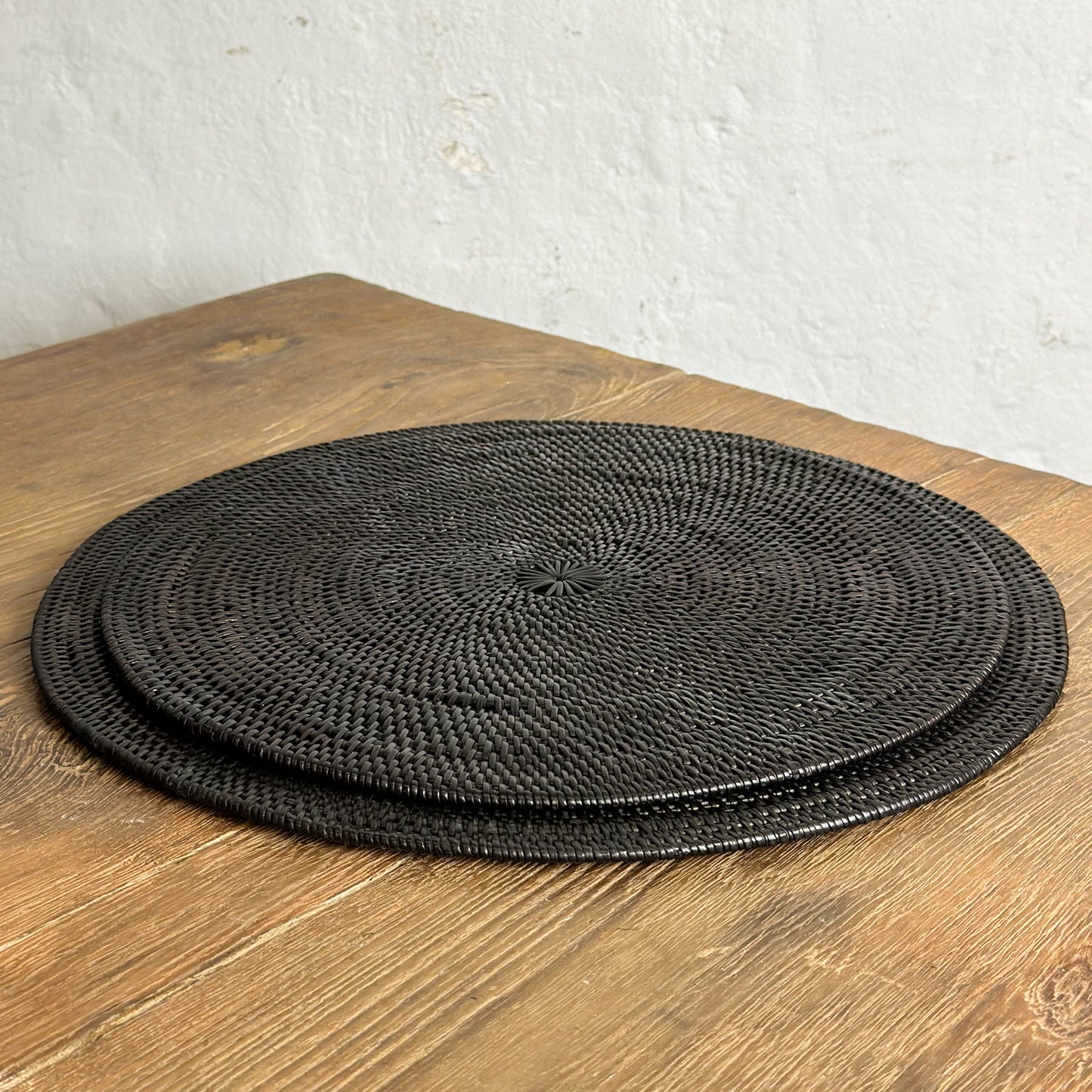 A stack of round black placemats on a wooden table surface against a white wall background.