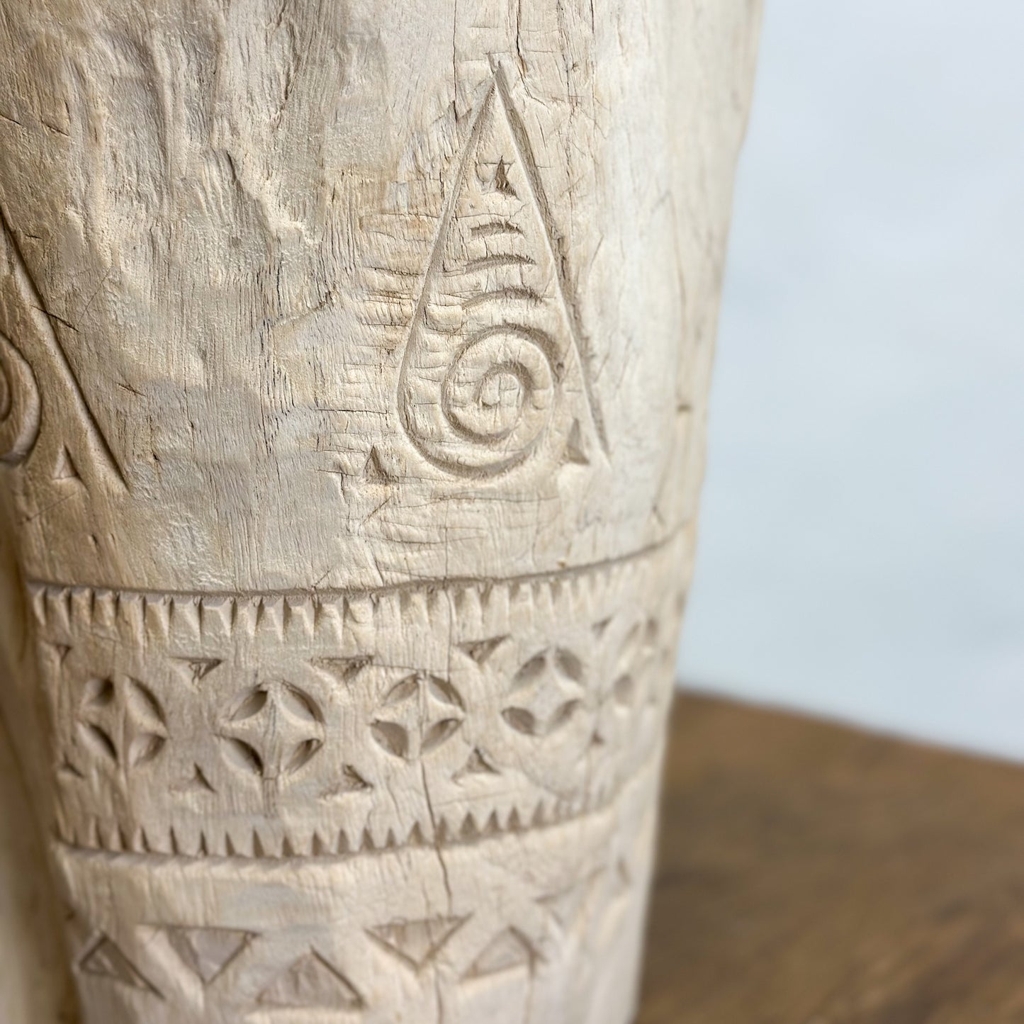 A bleached teak wood lesung (decorative vase) with carved patterns, placed on a wooden surface against a white wall.