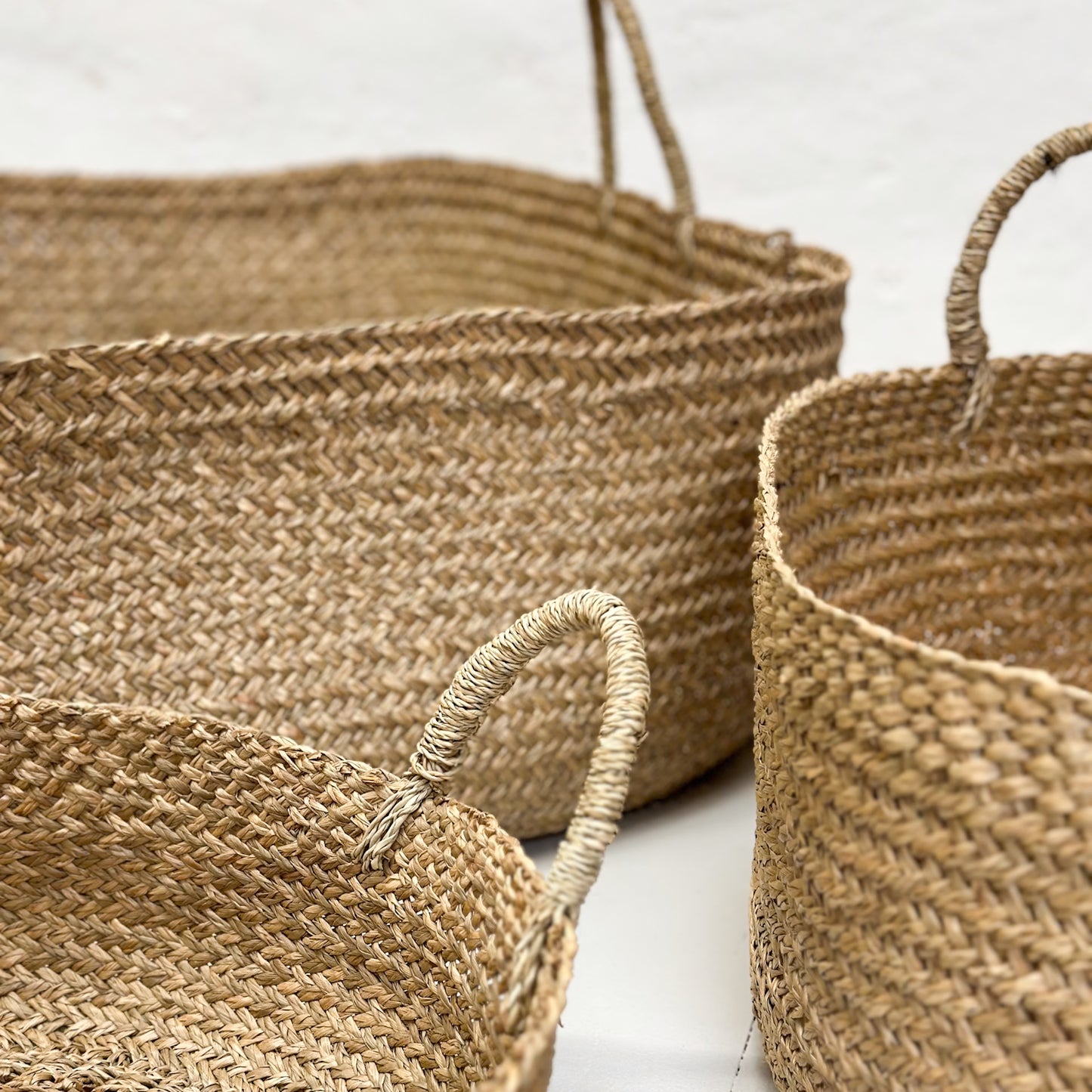 Three handcrafted wicker baskets in various sizes with natural sea grass color and texture, placed on a wooden floor against a white wall.