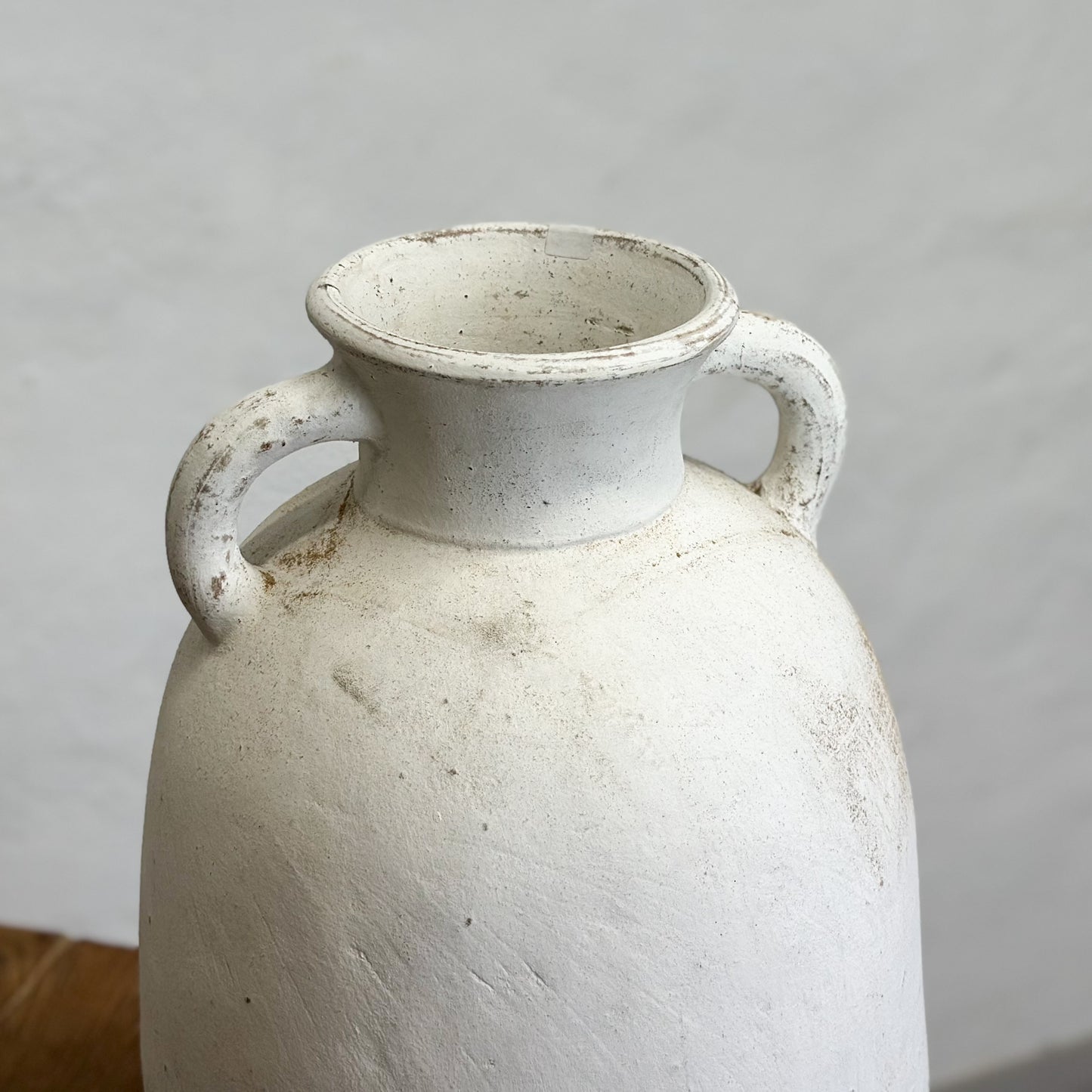 A terracotta pot with handles, positioned on a wooden surface against a plain wall.