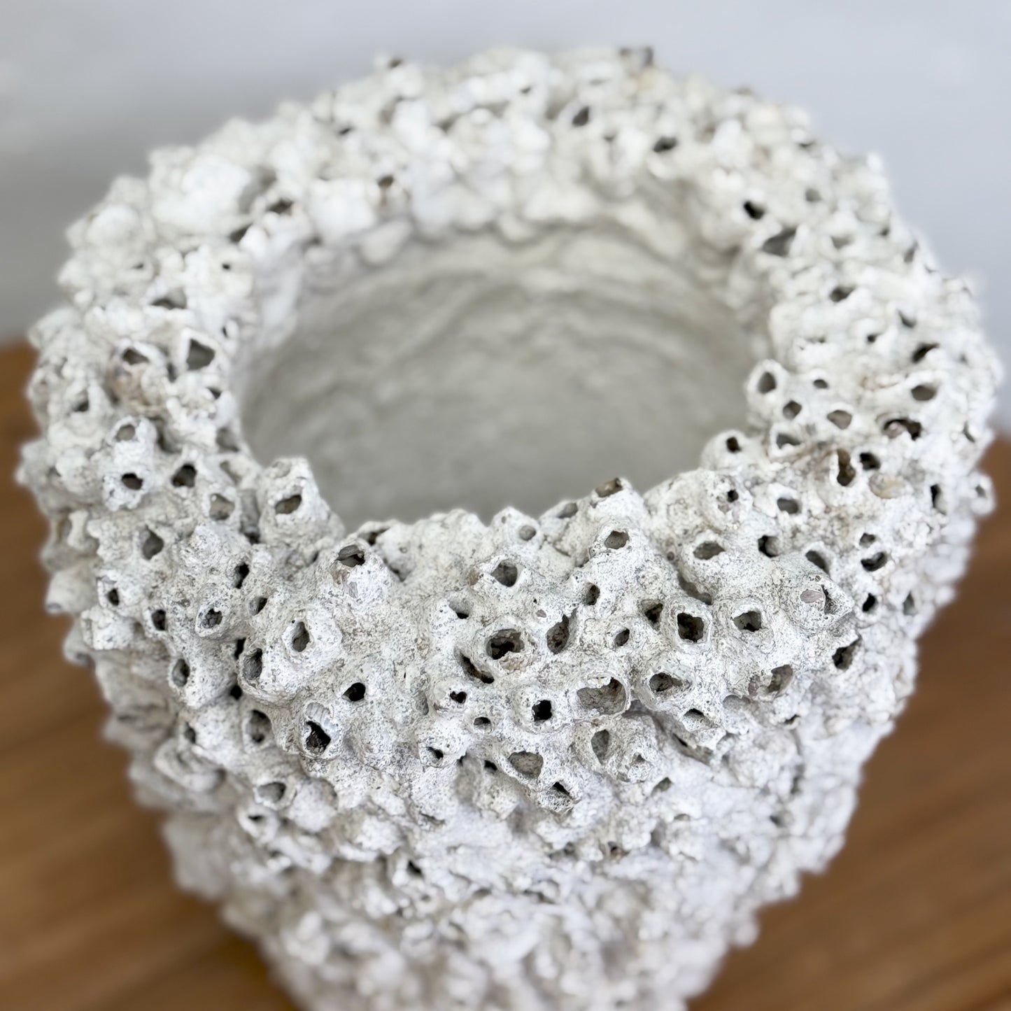 A beige ceramic pot with a textured surface, displayed on a wooden table against a plain wall.