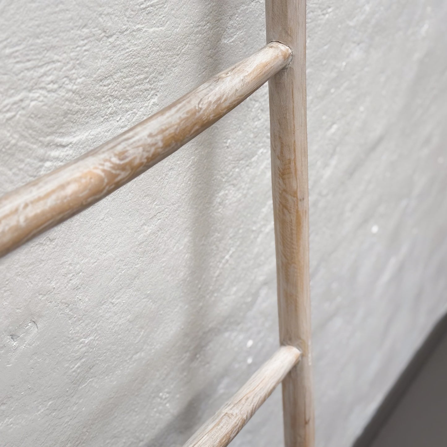 A beige suar wood ladder with a simple design, leaning against a plain wall on a grey floor.