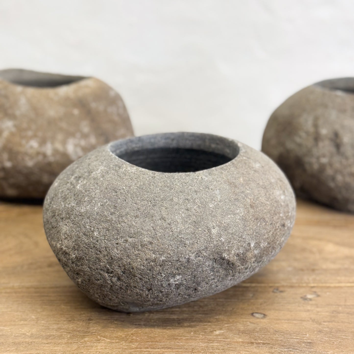 Three Balinese sculpted stone bowls of varying sizes displayed on a wooden surface against a white wall.