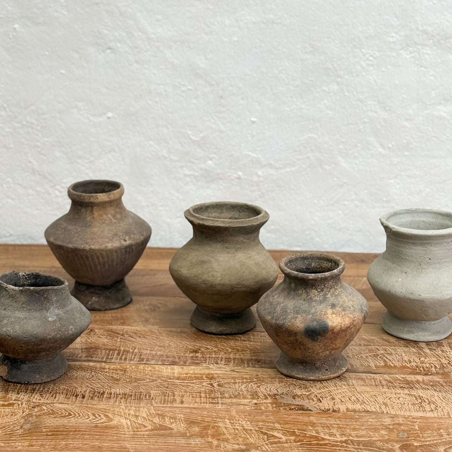 Five rustic clay pots of varying sizes on a wooden surface with a textured white wall background.