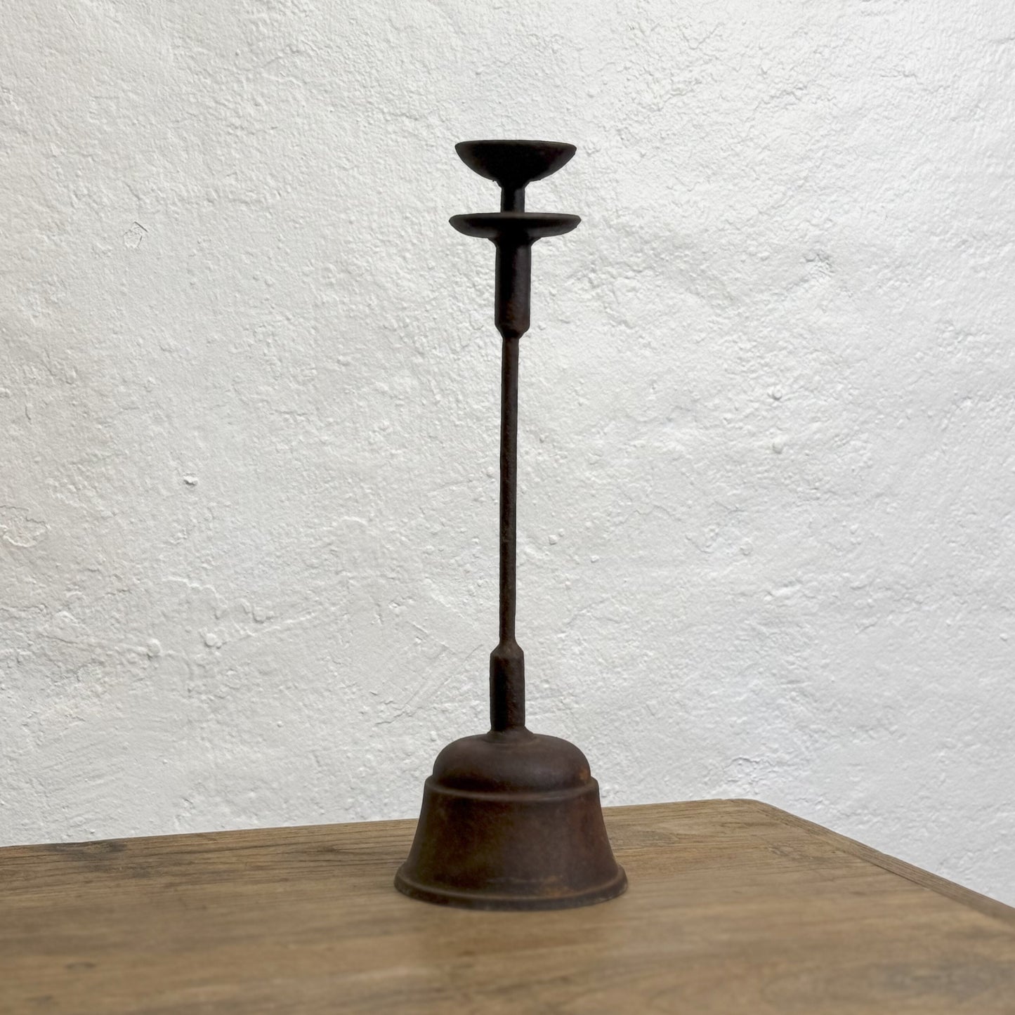 A vintage iron oil lamp with a cylindrical base and a tapered oil container, placed on a wooden surface against a white wall.