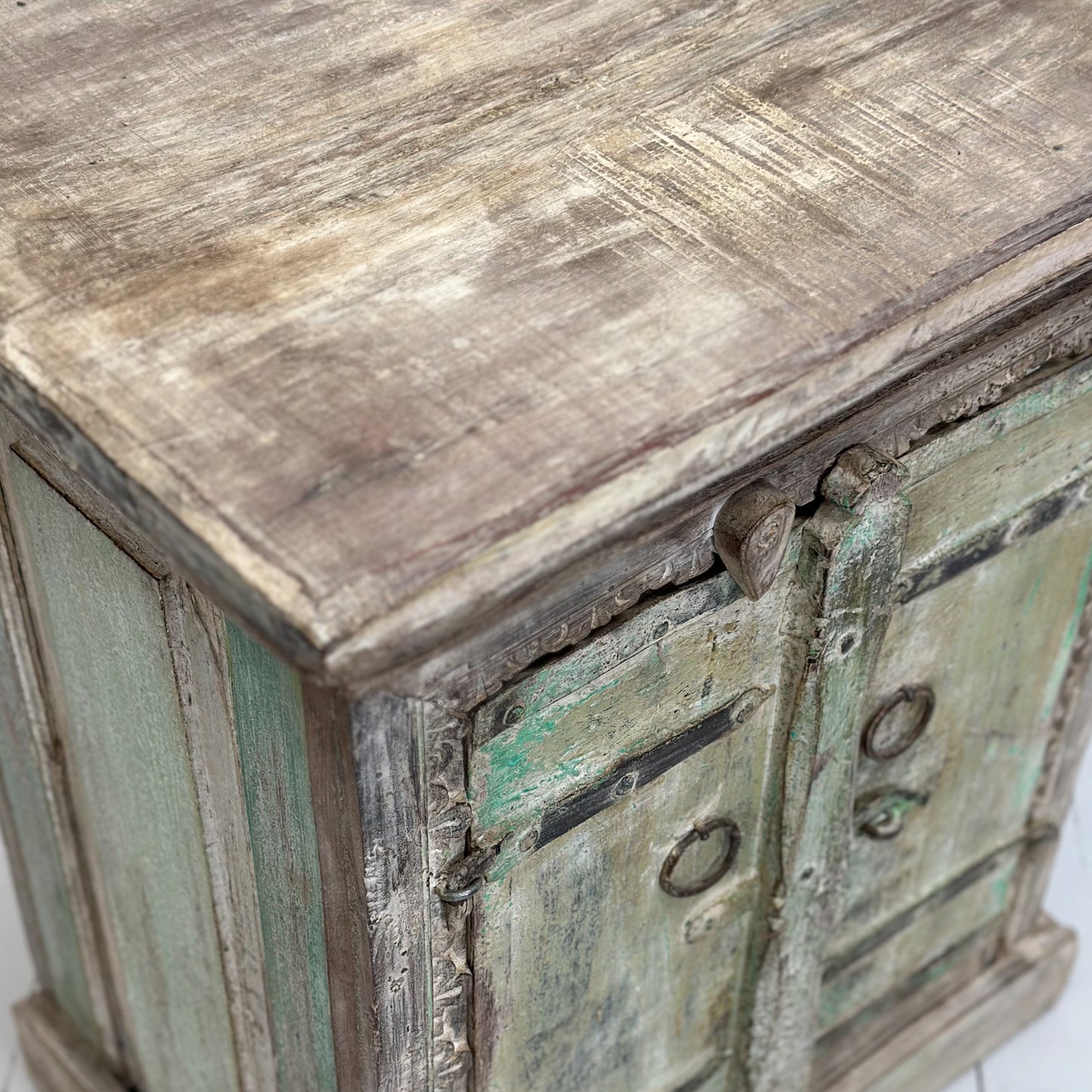 A carved wooden bedside cabinet with a green patina on the doors and a beige textured surface, placed against a plain wall on a white floor.