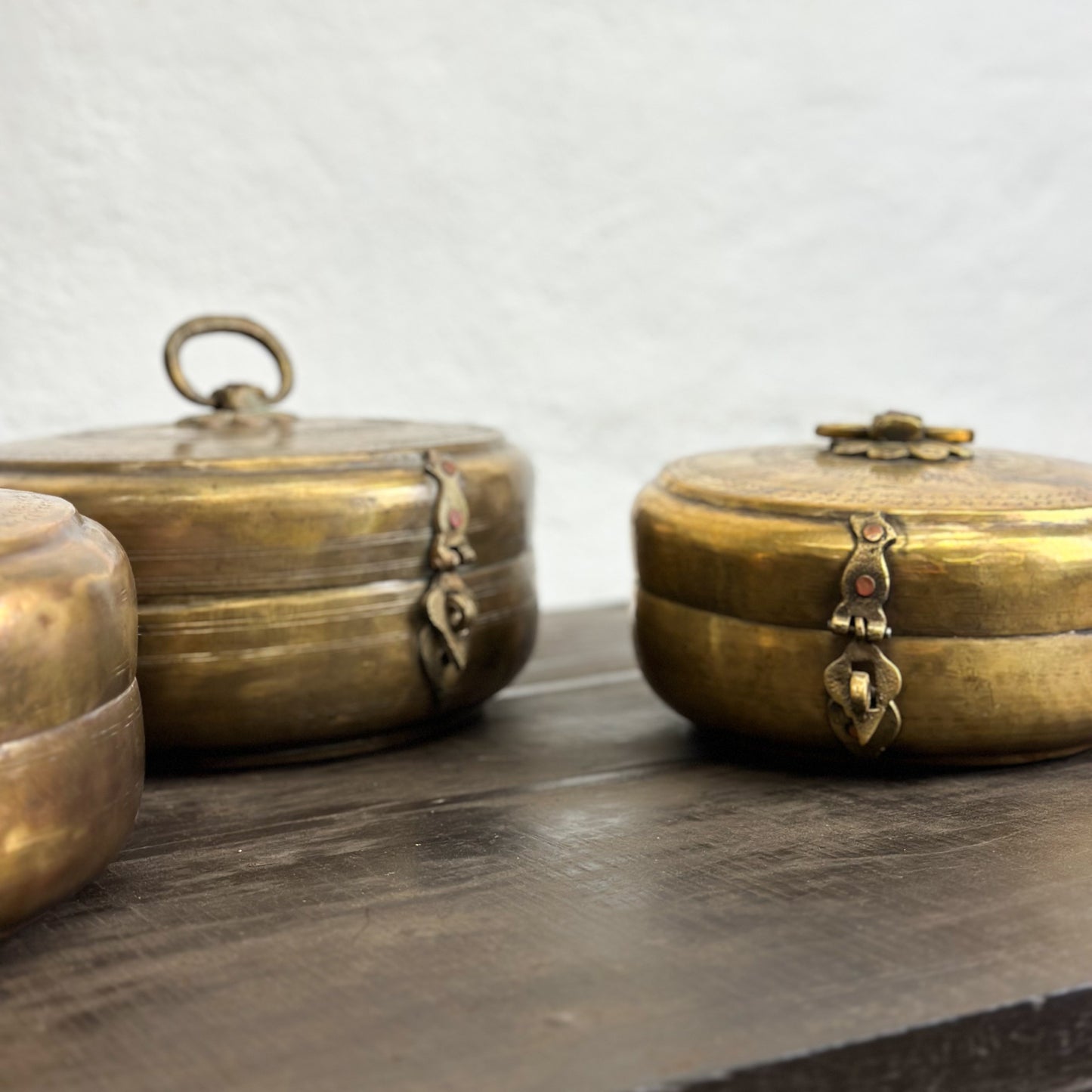 Three brass chapati boxes with lids, placed on a wooden surface against a white wall background.