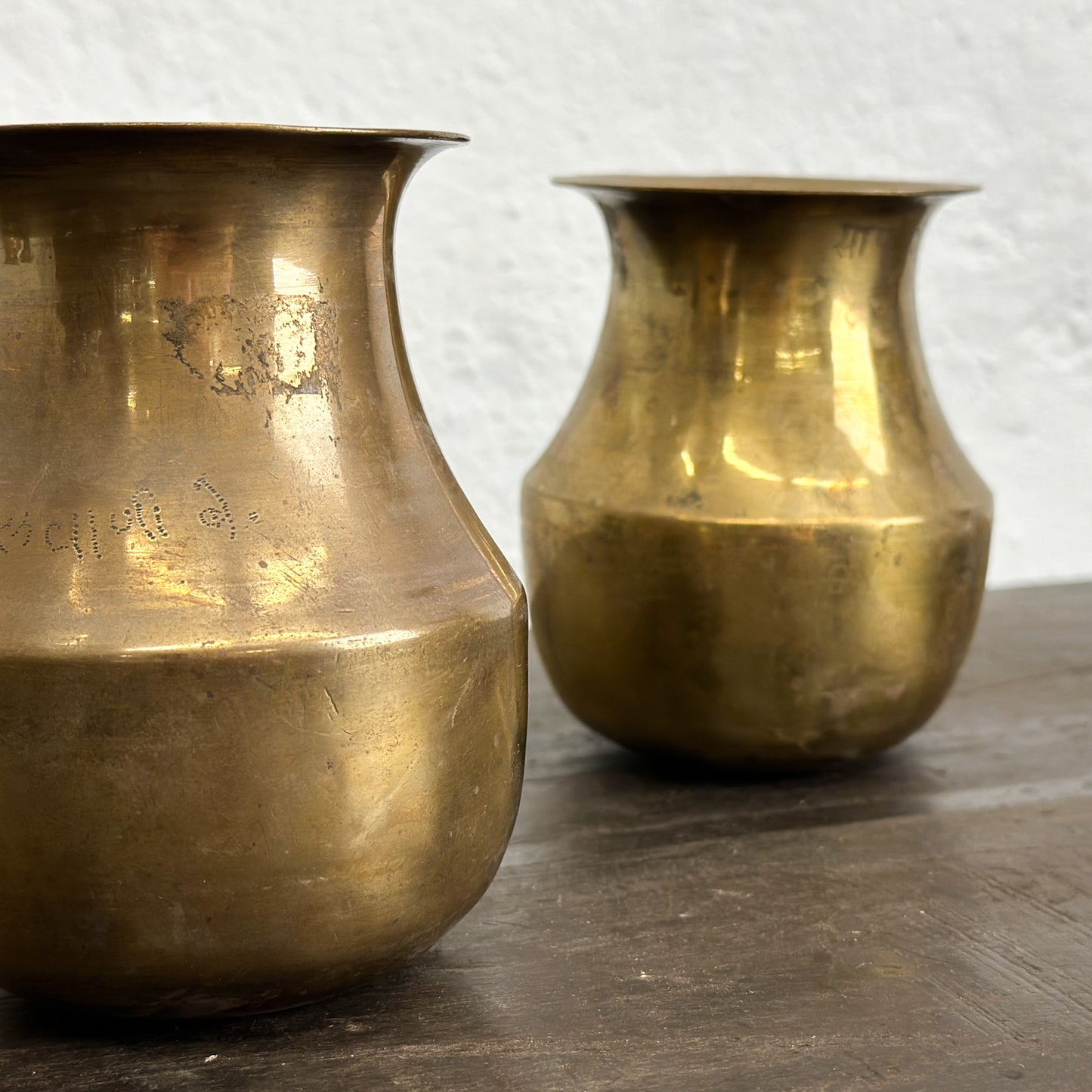Two brass watering cans placed side by side on a wooden surface against a white wall background.