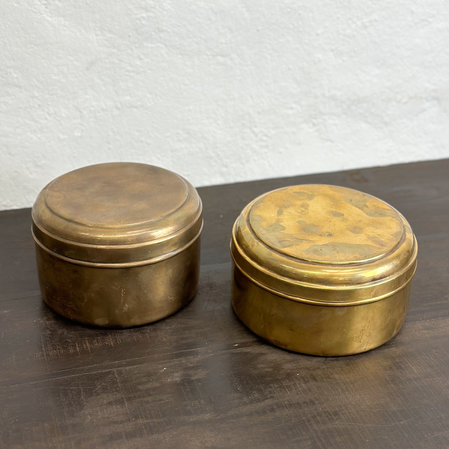 Two brass lunch boxes with a lid, one on top of the other, placed on a wooden surface against a white wall.