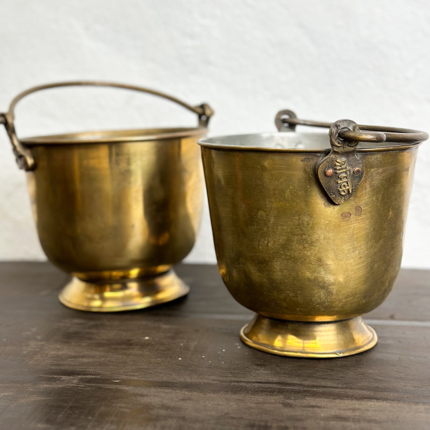 Three small brass buckets with bases, two with handles, placed on a wooden surface against a white wall.