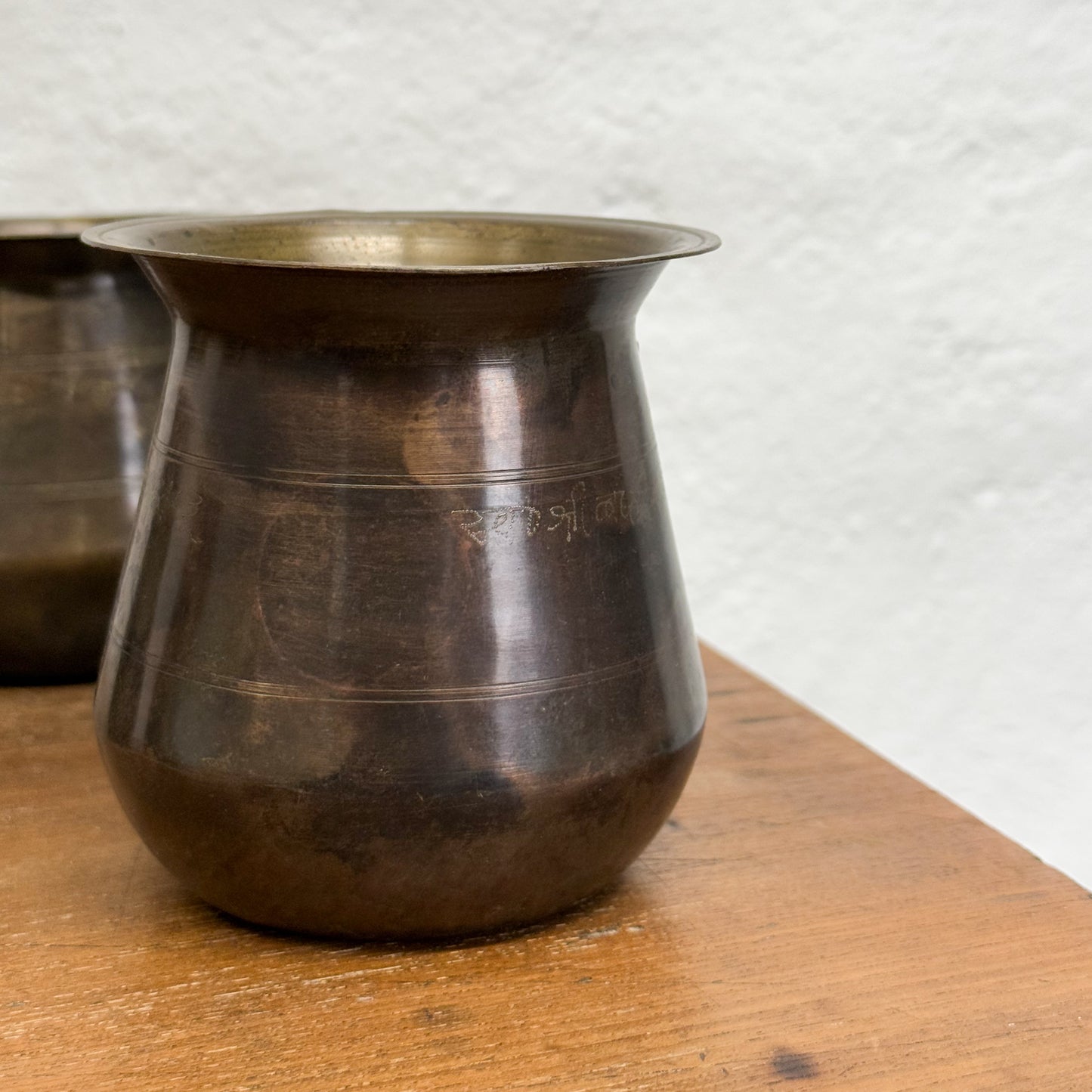 A collection of vintage Indian brass Lota displayed on a wooden surface against a white wall.