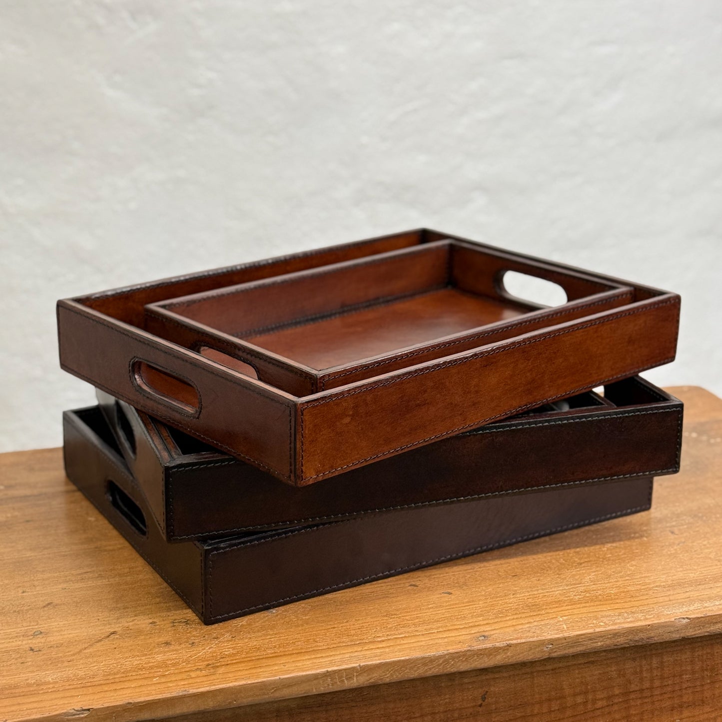 Two brown leather trays with handles, one fits inside the other, on a wooden surface.