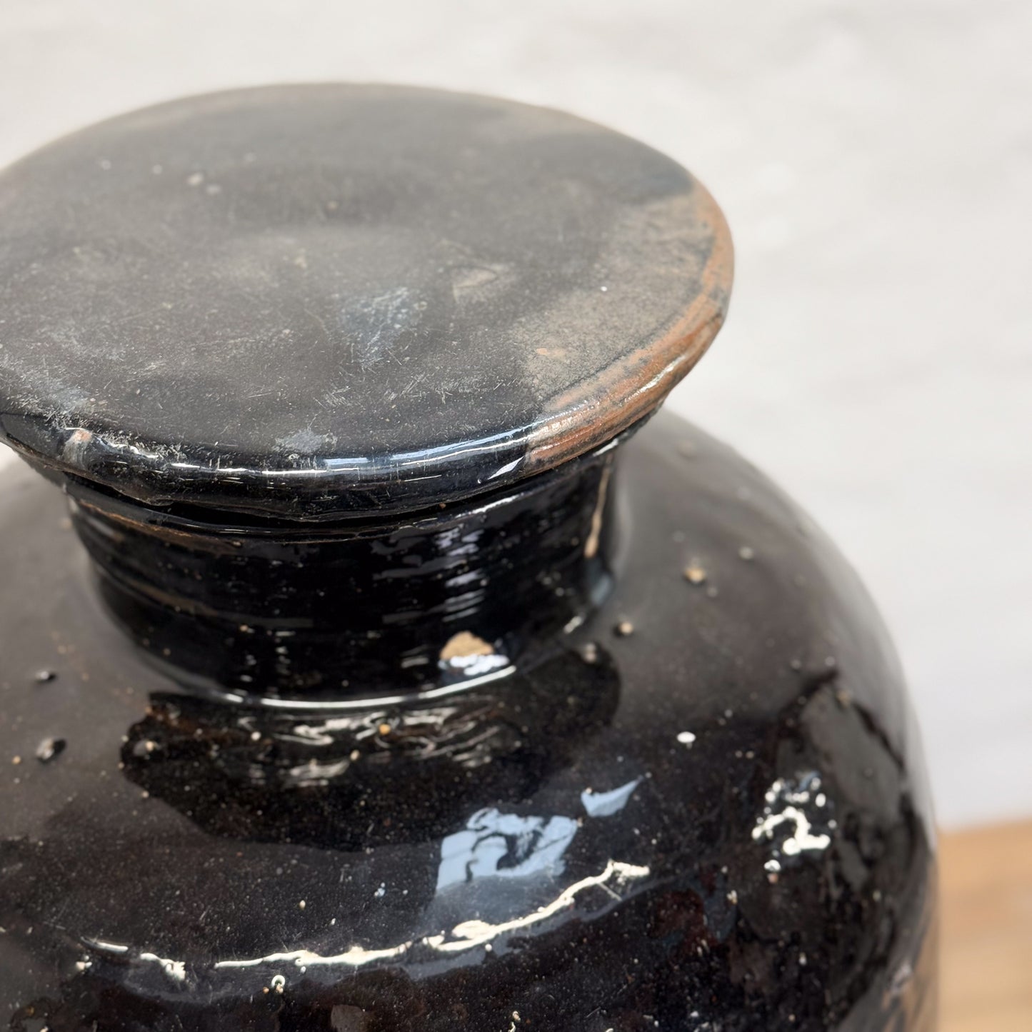A black, glazed, lidded storage jar placed on a wooden surface with a plain wall in the background