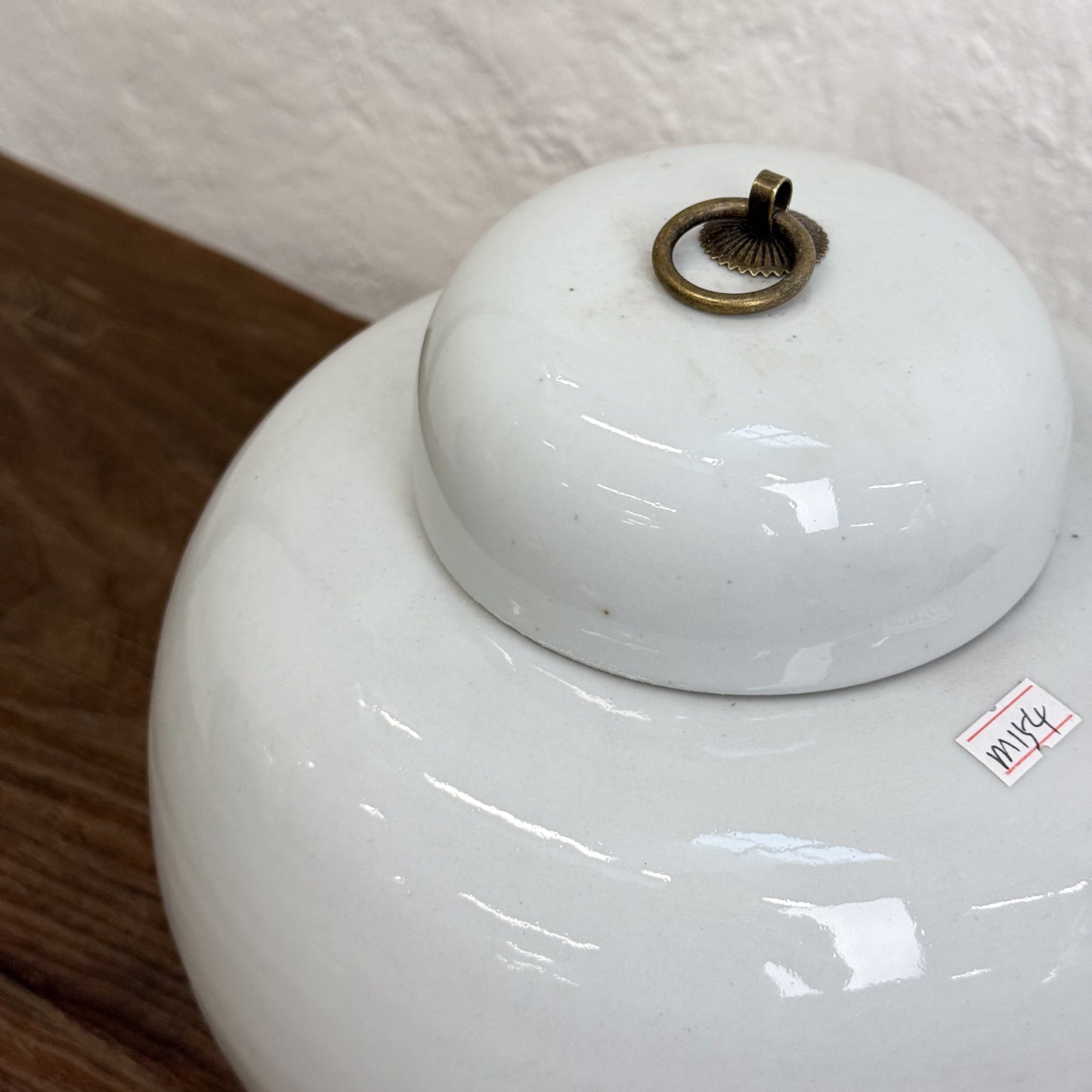 A white, porcelain, squat melon jar with a lid on top, displayed on a wooden surface against a white background.