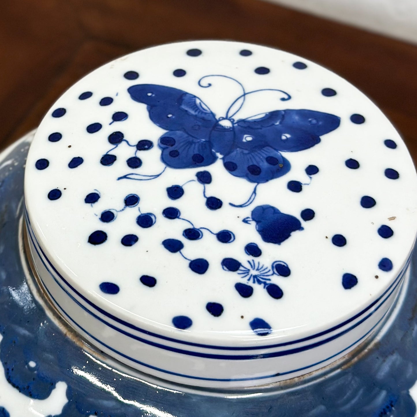 A blue porcelain ginger jar with a butterfly pattern and dotted details, displayed on a wooden surface against a textured background.