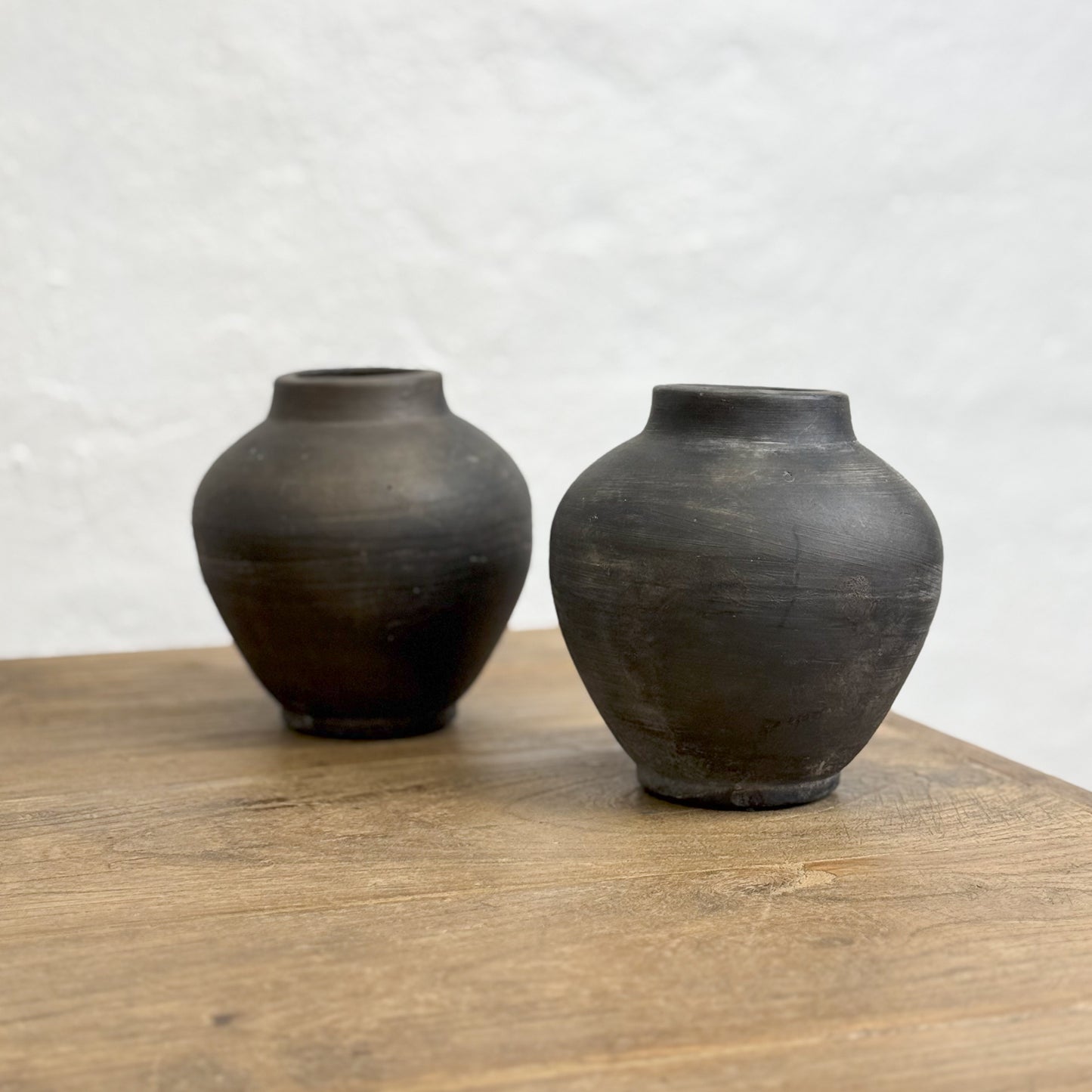 A small earthenware vase with a matte black finish, placed on a wooden surface against a textured wall.