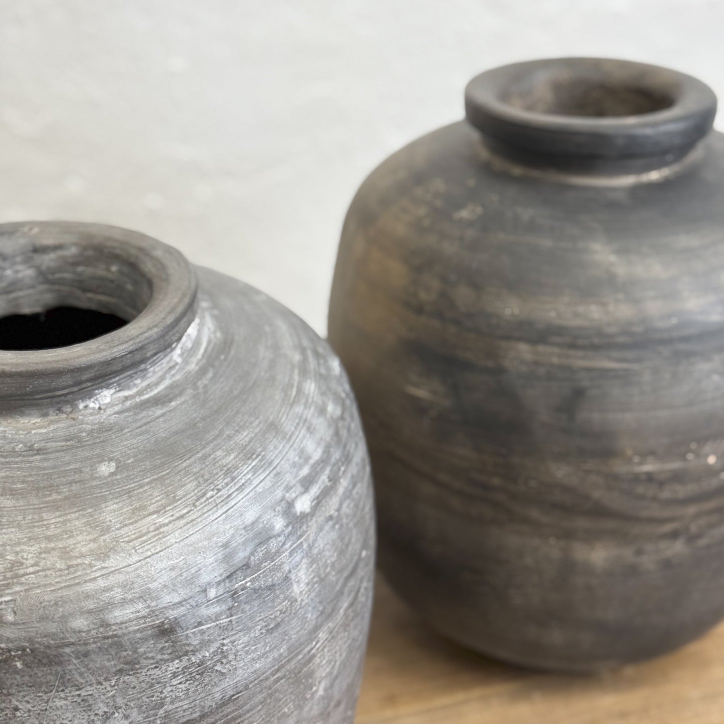 A large earthenware vase with a lip, featuring a dark gray or black color and a textured surface, placed on a wooden surface against a white wall