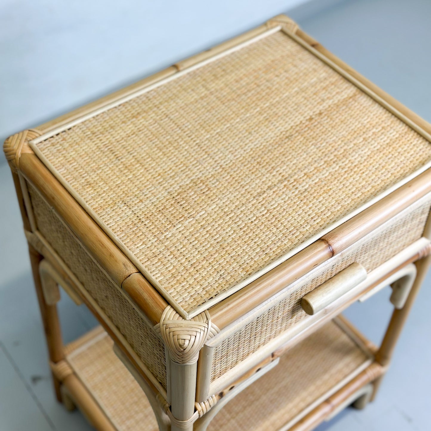 A beige cane and rattan bedside table with a drawer and a lower shelf, placed against a neutral background.
