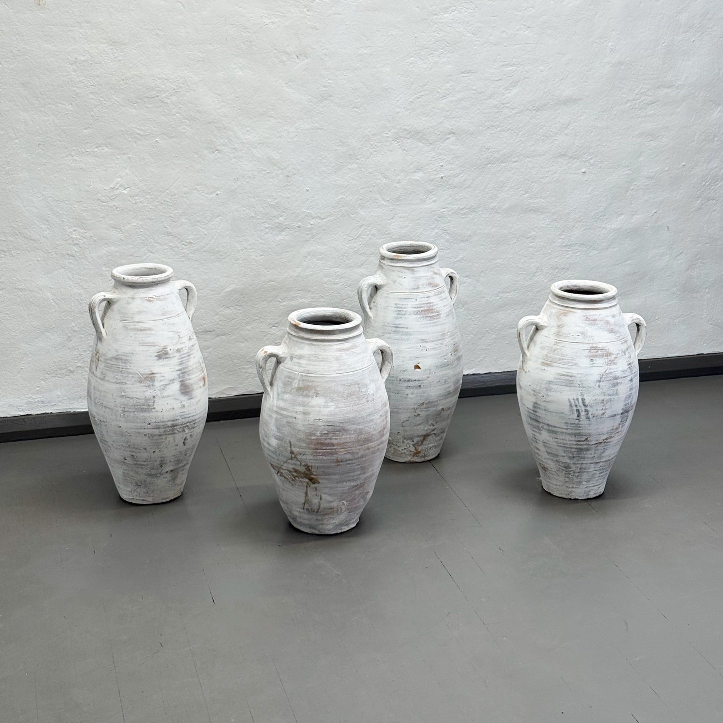 Four white ceramic vases with handles on a gray floor against a plain wall.