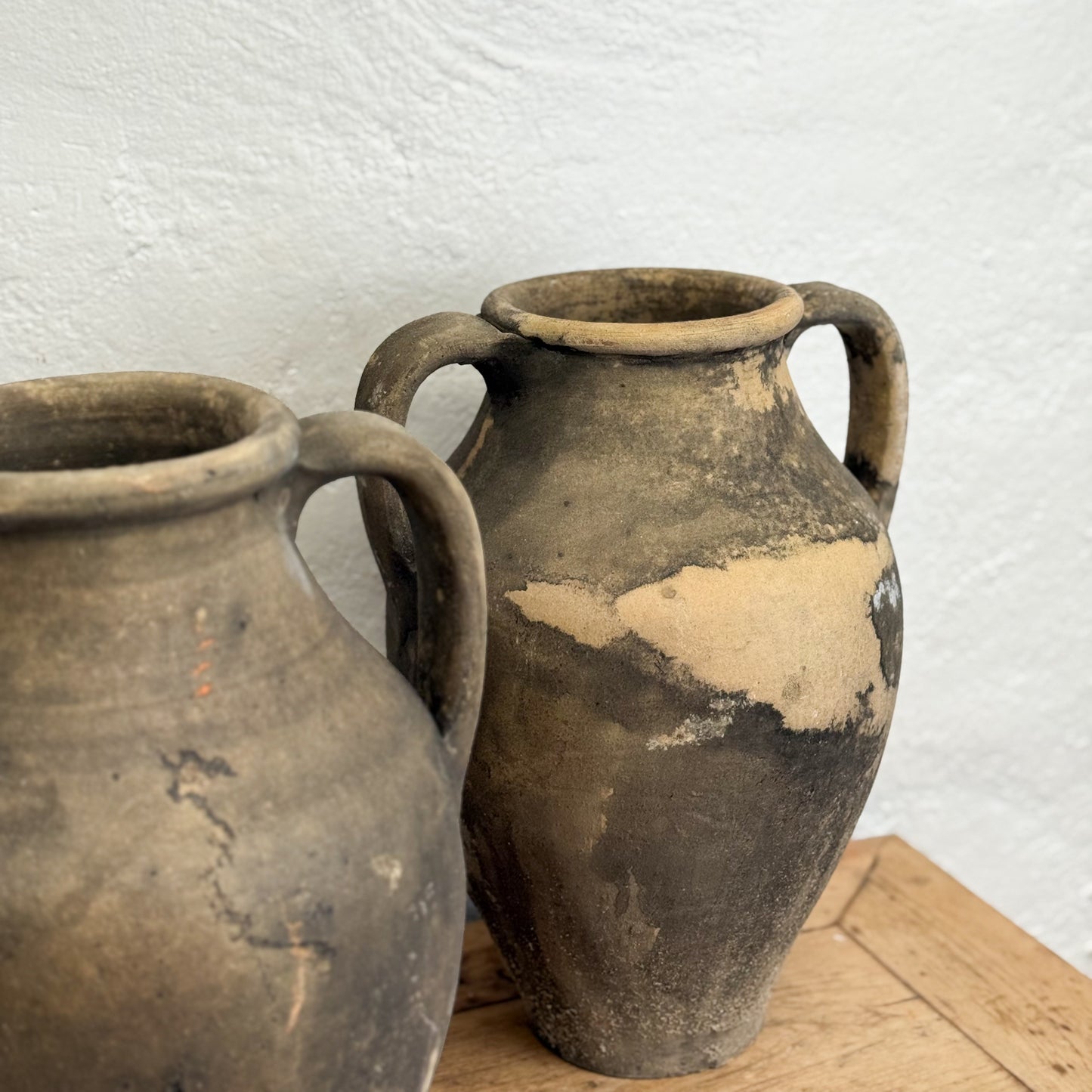 Two rustic clay pots with handles on a wooden surface against a white wall.