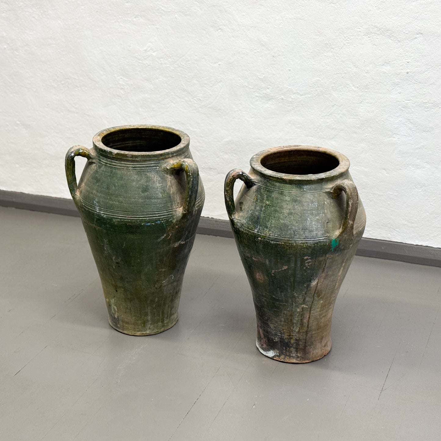 Two green ceramic vases with handles on a light gray floor against a white textured wall.