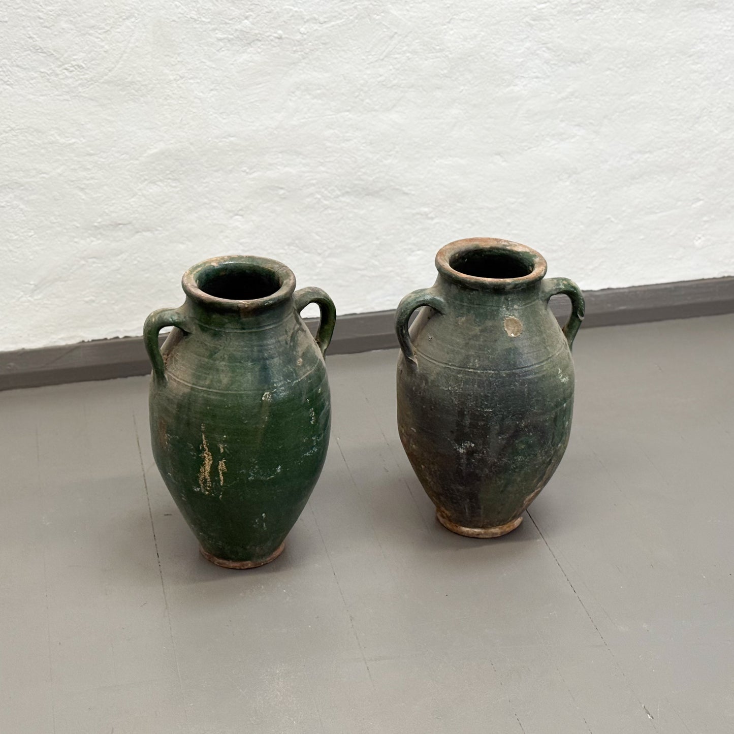 Two green ceramic vases on a light gray floor with a white textured wall in the background.