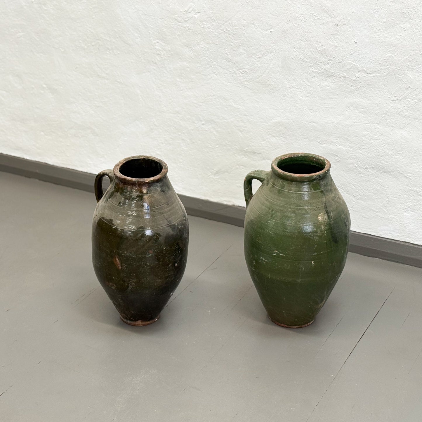 Two ceramic vases on a light gray floor with a textured white wall in the background.