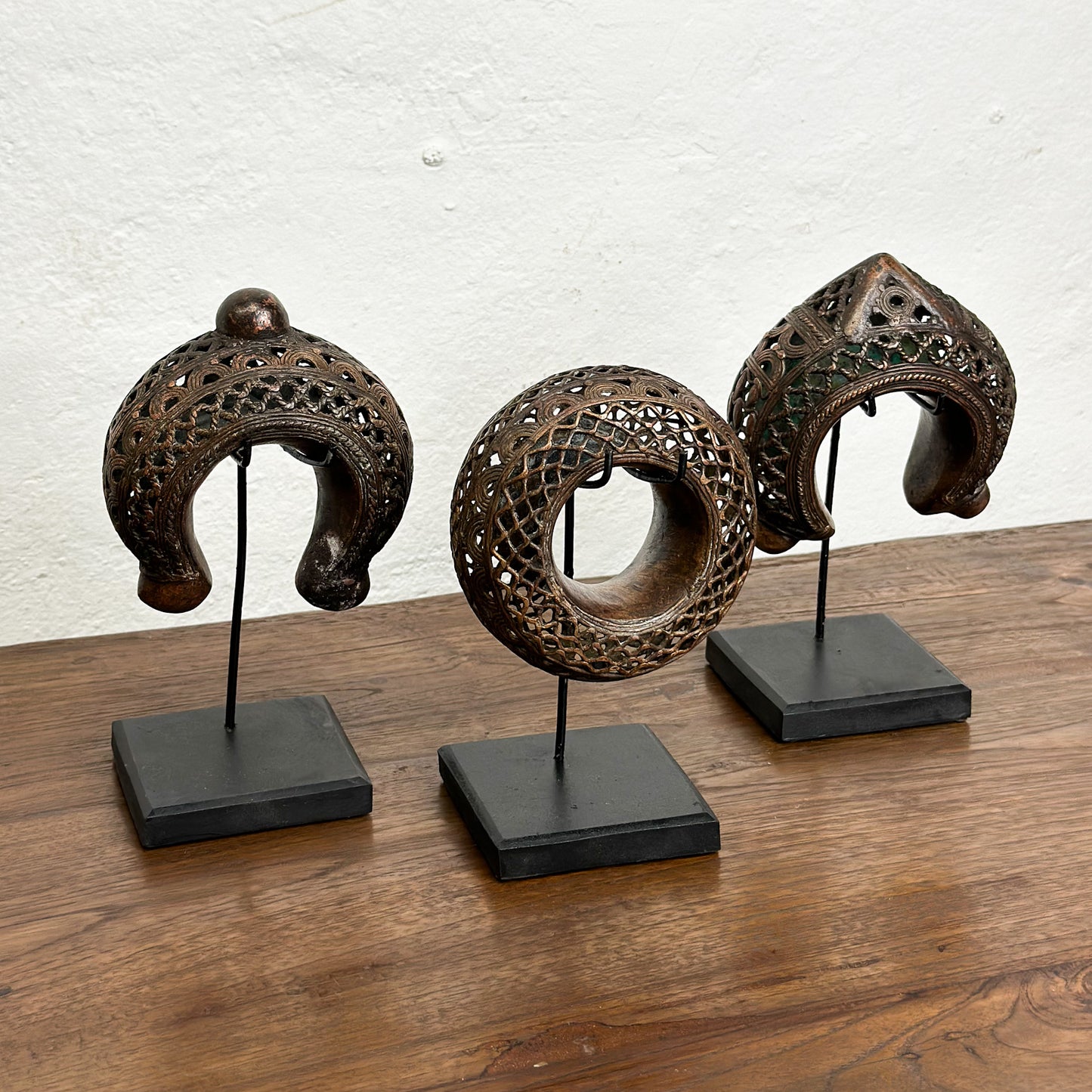 Three bronze anklets with intricate geometric patterns displayed on individual stands against a white background.