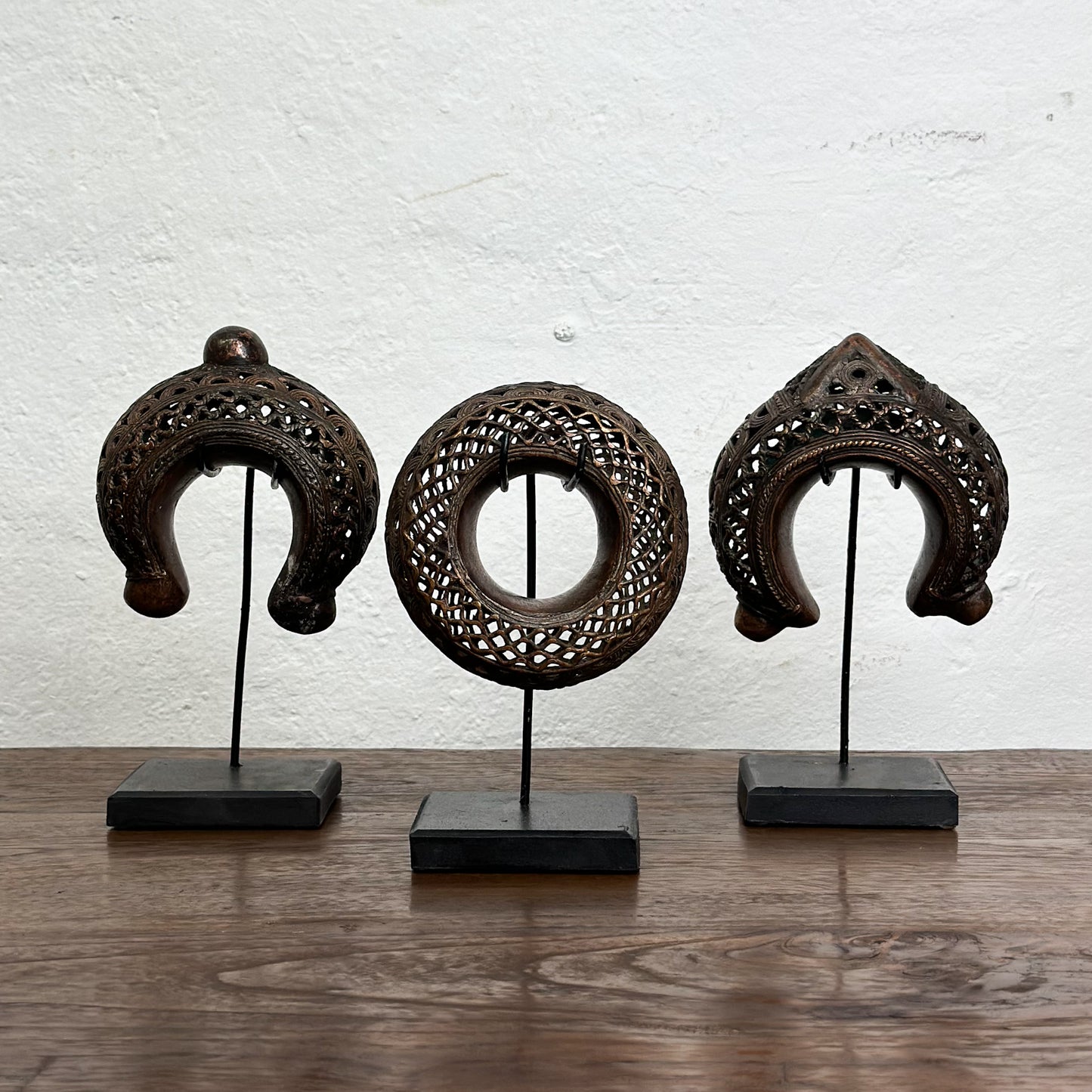 Three bronze anklets with intricate geometric patterns displayed on individual stands against a white background.