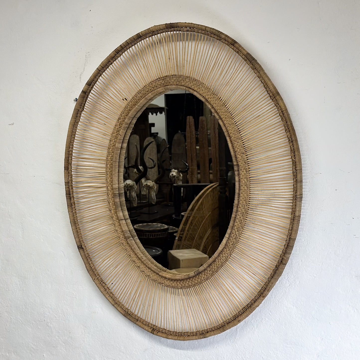 An oval-shaped mirror with a natural, woven frame, reflecting a room with white walls and a wooden chair.