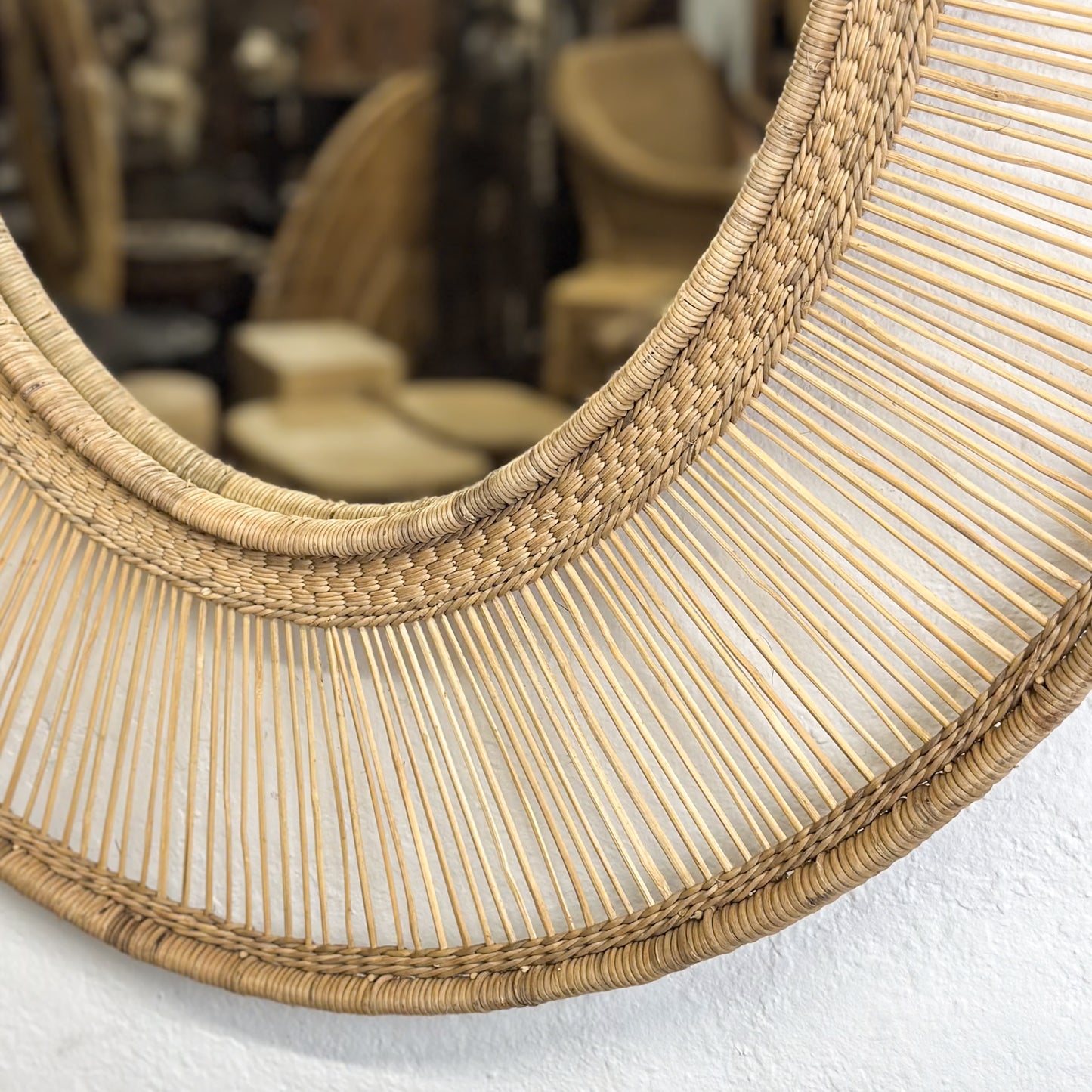 An oval-shaped mirror with a natural, woven frame, reflecting a room with white walls and a wooden chair.