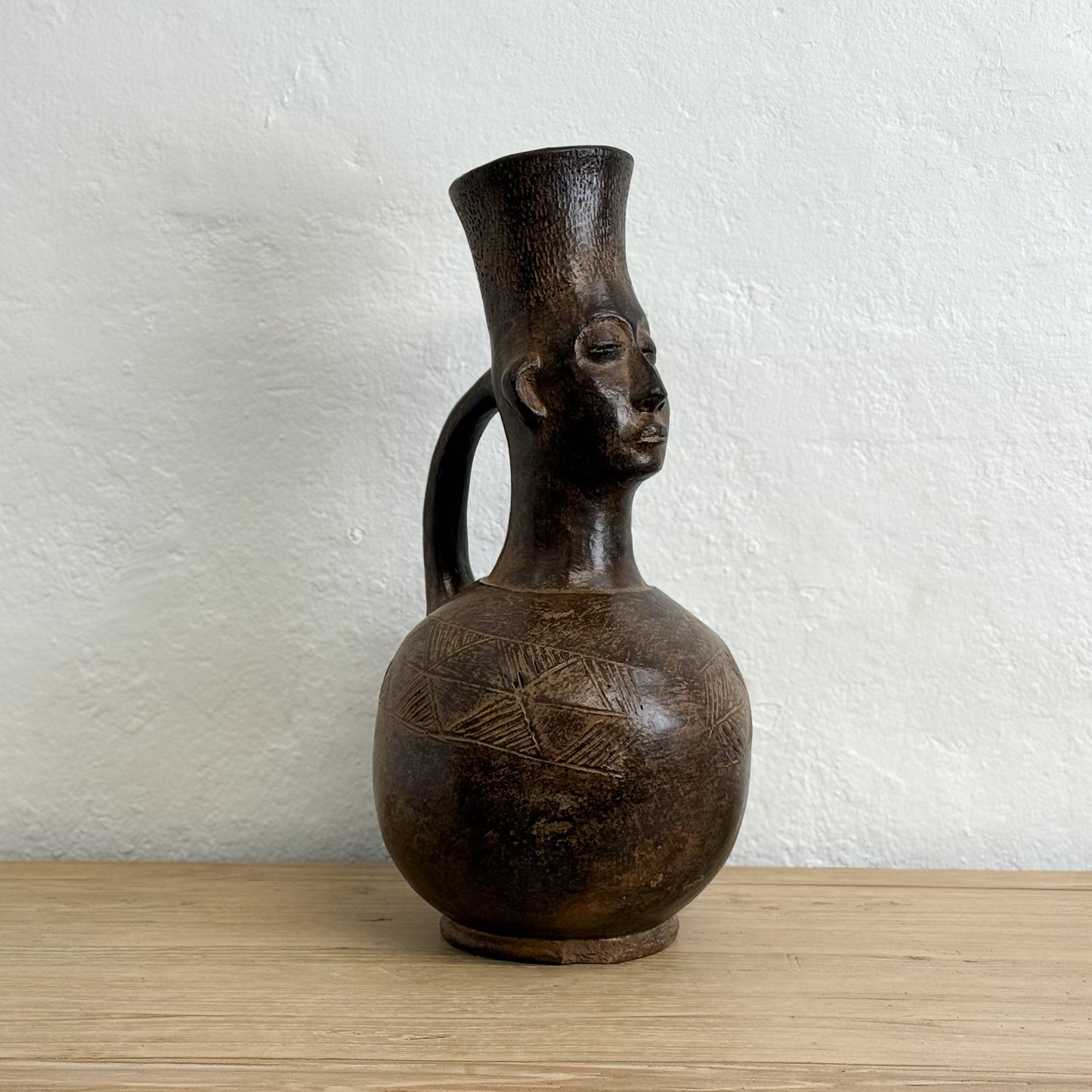 Brown ceramic vase with a face design on a wooden surface and white textured wall background