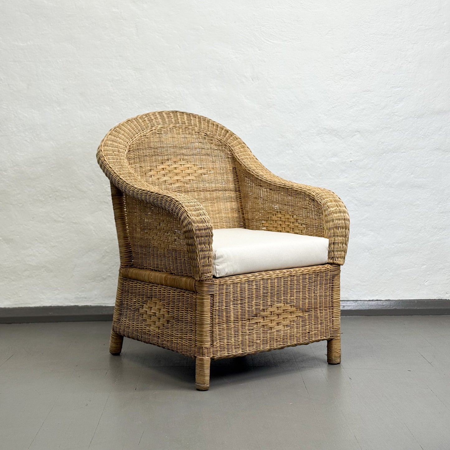 Wicker armchair with a white cushion against a plain wall.