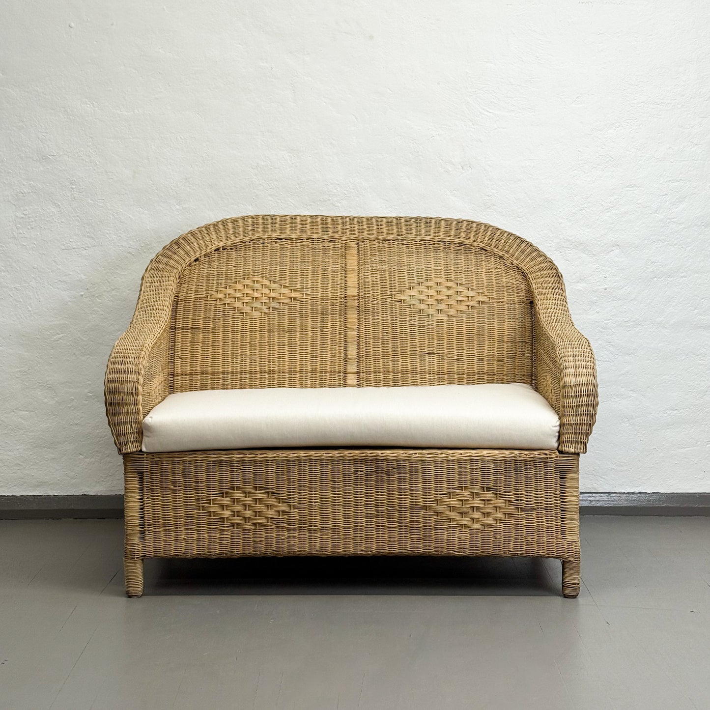 Wicker chair with a white cushion against a plain wall.