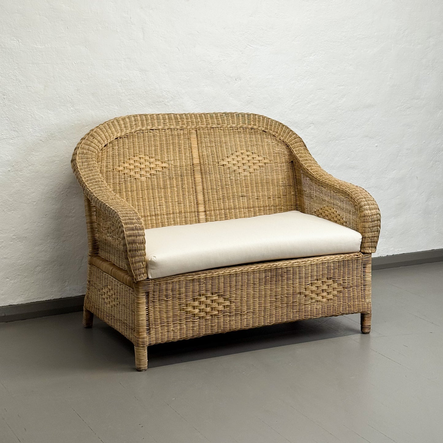 Wicker loveseat with a white cushion against a plain wall.