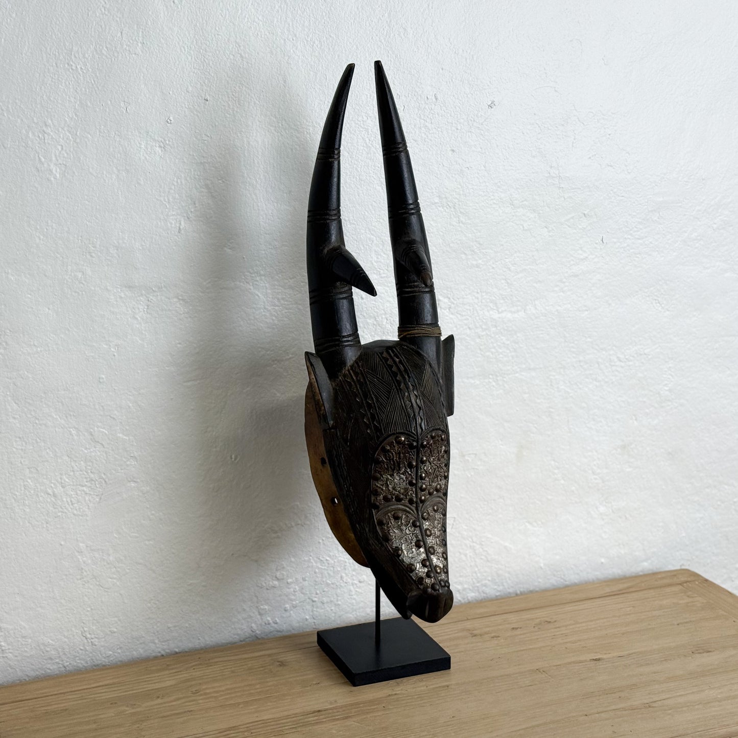 Decorative mask with large horns on a stand against a white wall.
