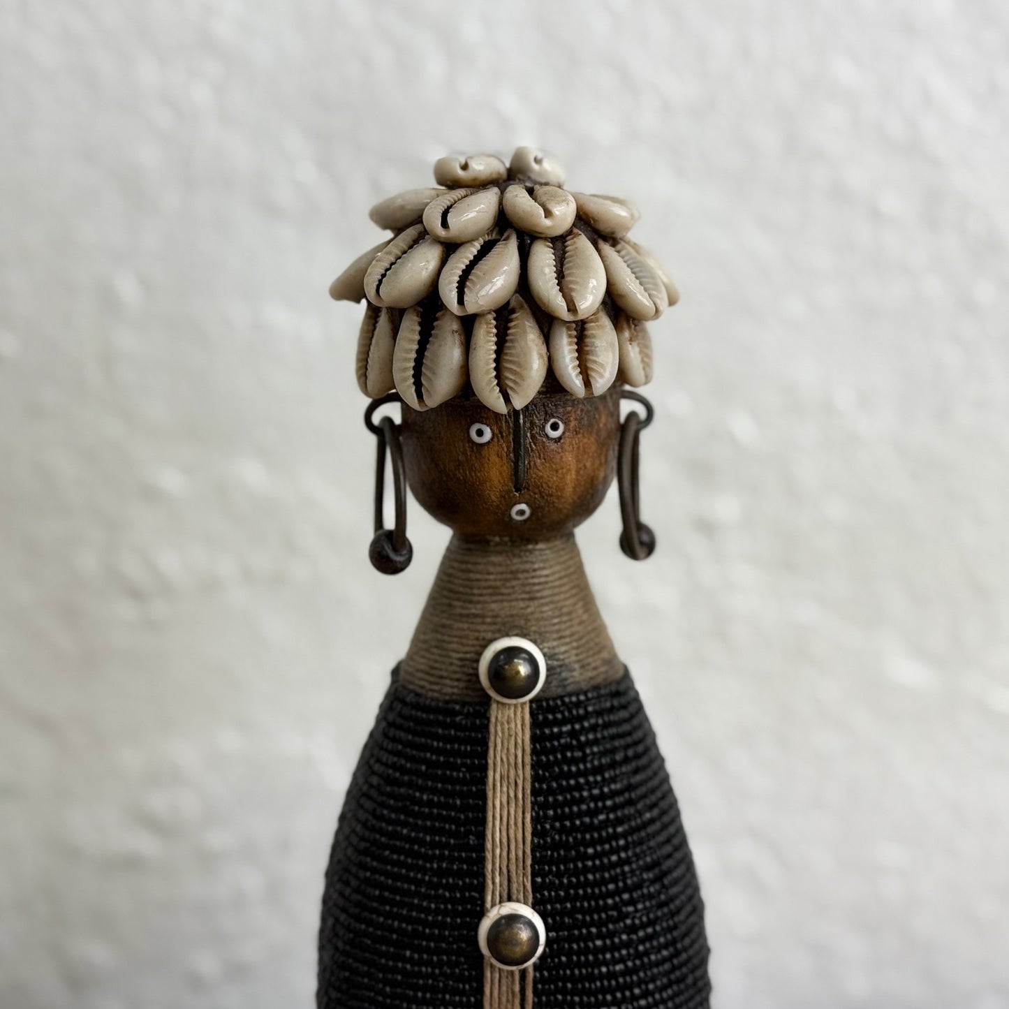 Decorative figure with shell headpiece and braided design on a textured white background