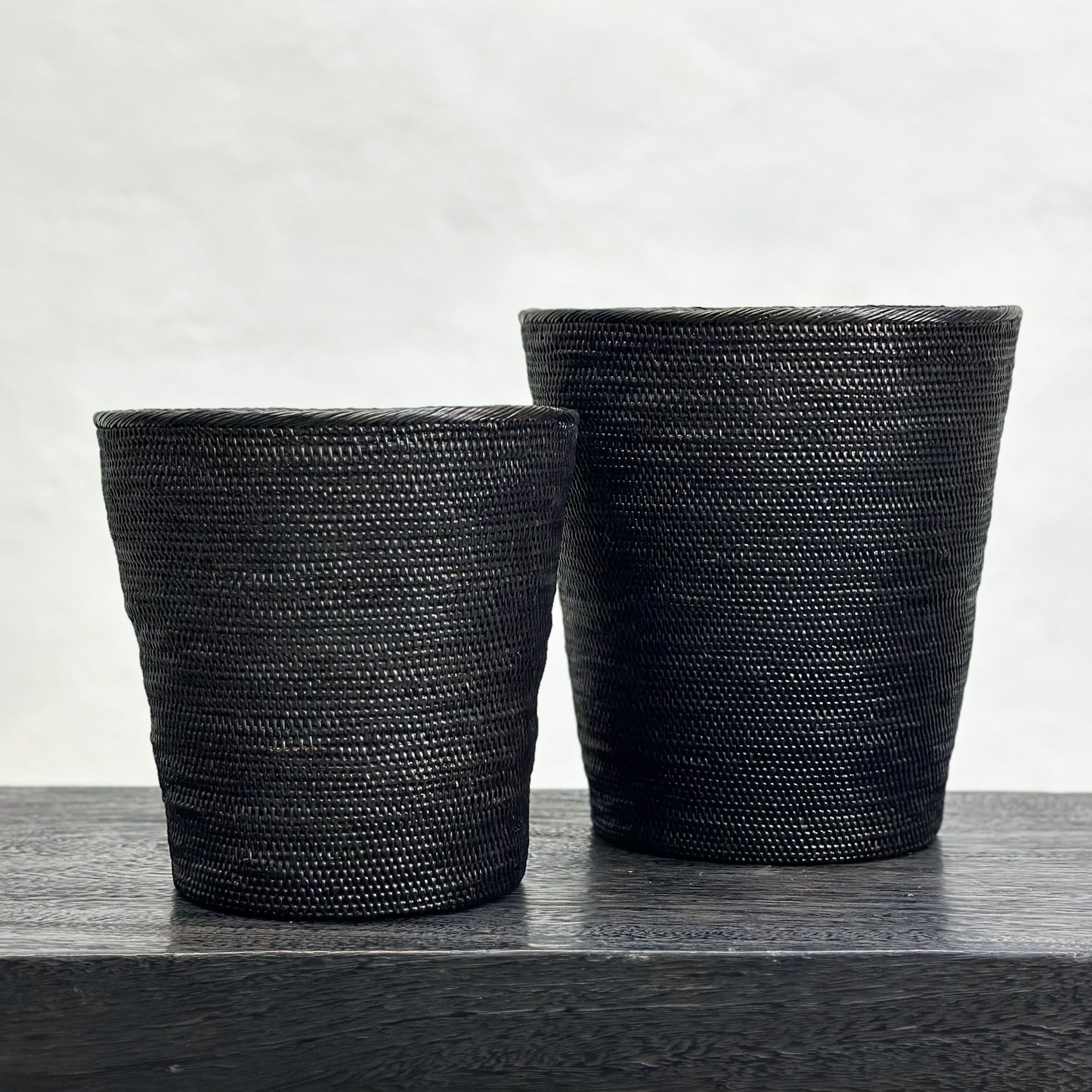 Two black tapered dustbins with a woven texture, placed on a dark surface.