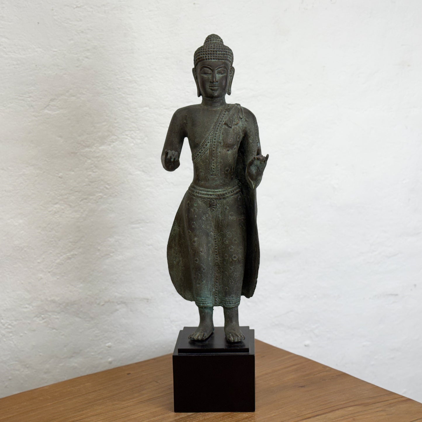 A bronze sculpture of a standing Buddha with hands outstretched, wearing a robe, placed on a black base.