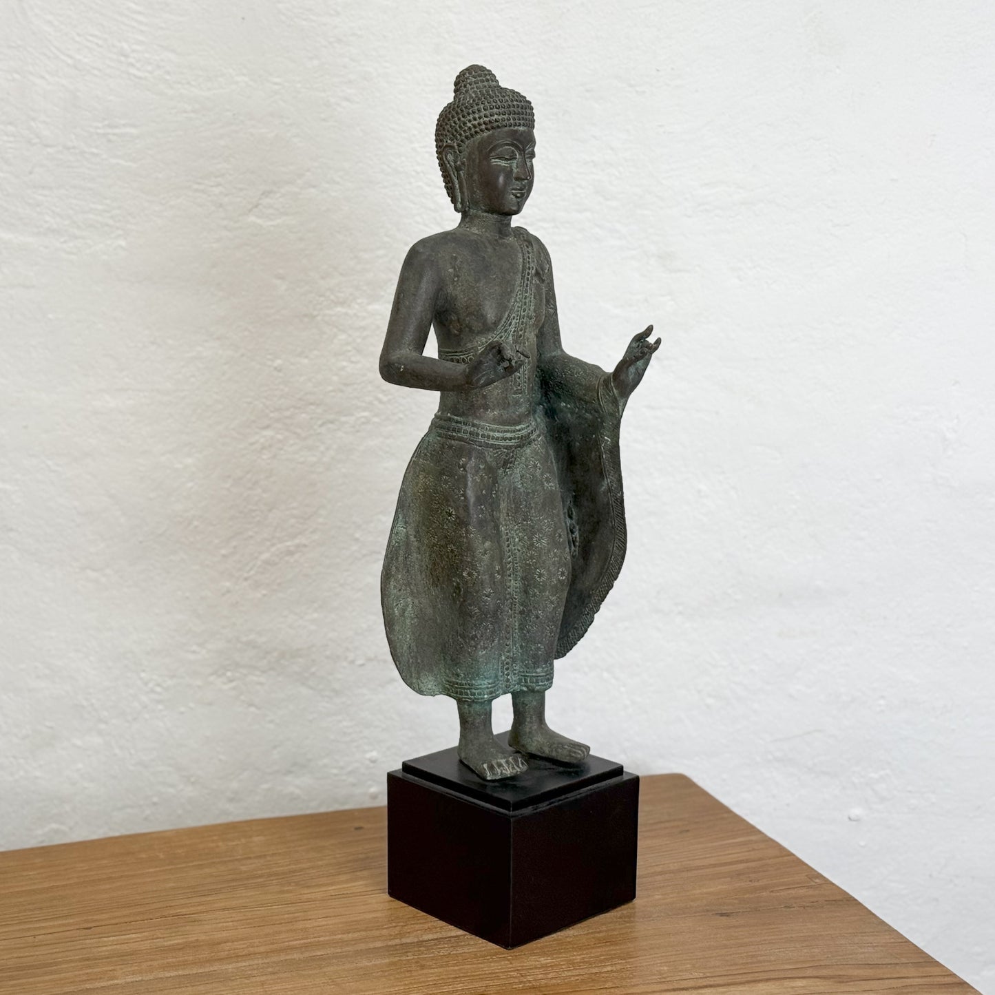 A bronze sculpture of a standing Buddha with hands outstretched, wearing a robe, placed on a black base.