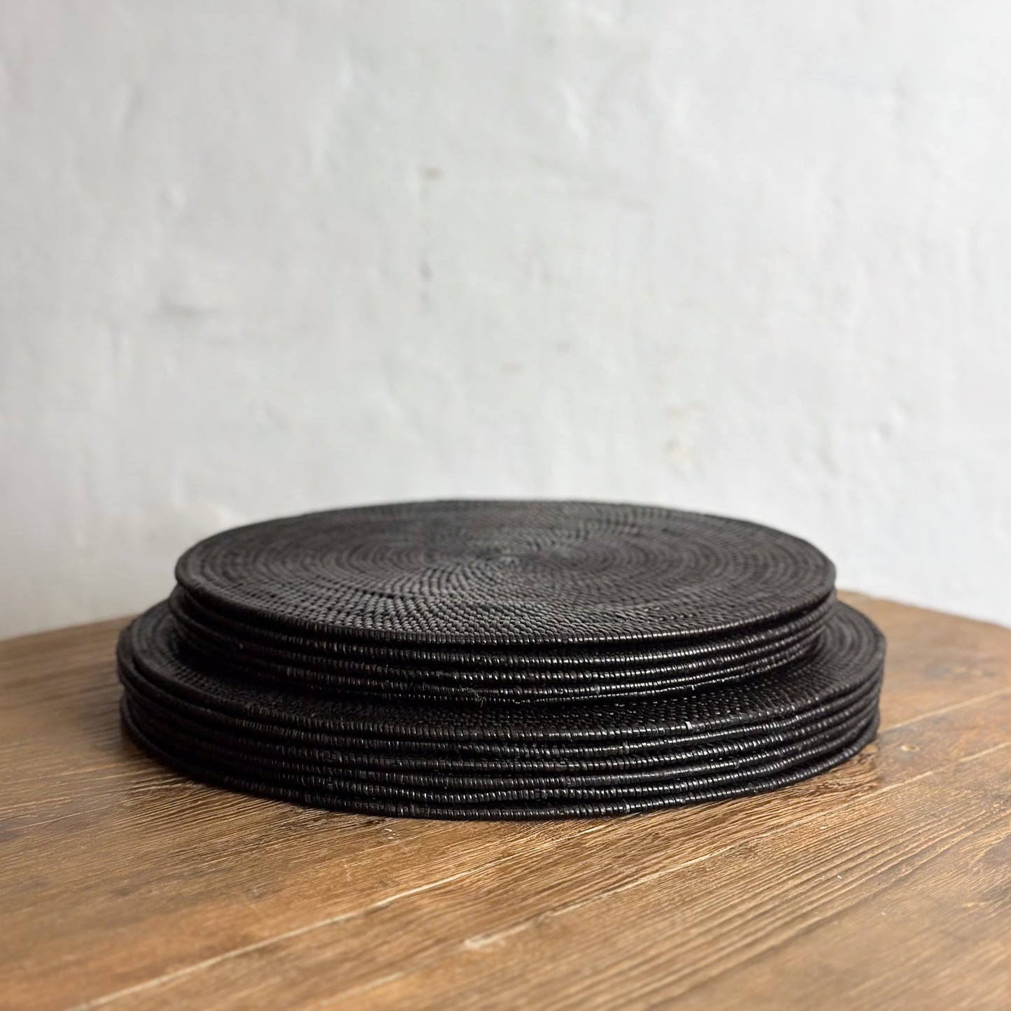 A stack of round black placemats on a wooden table surface against a white wall background.