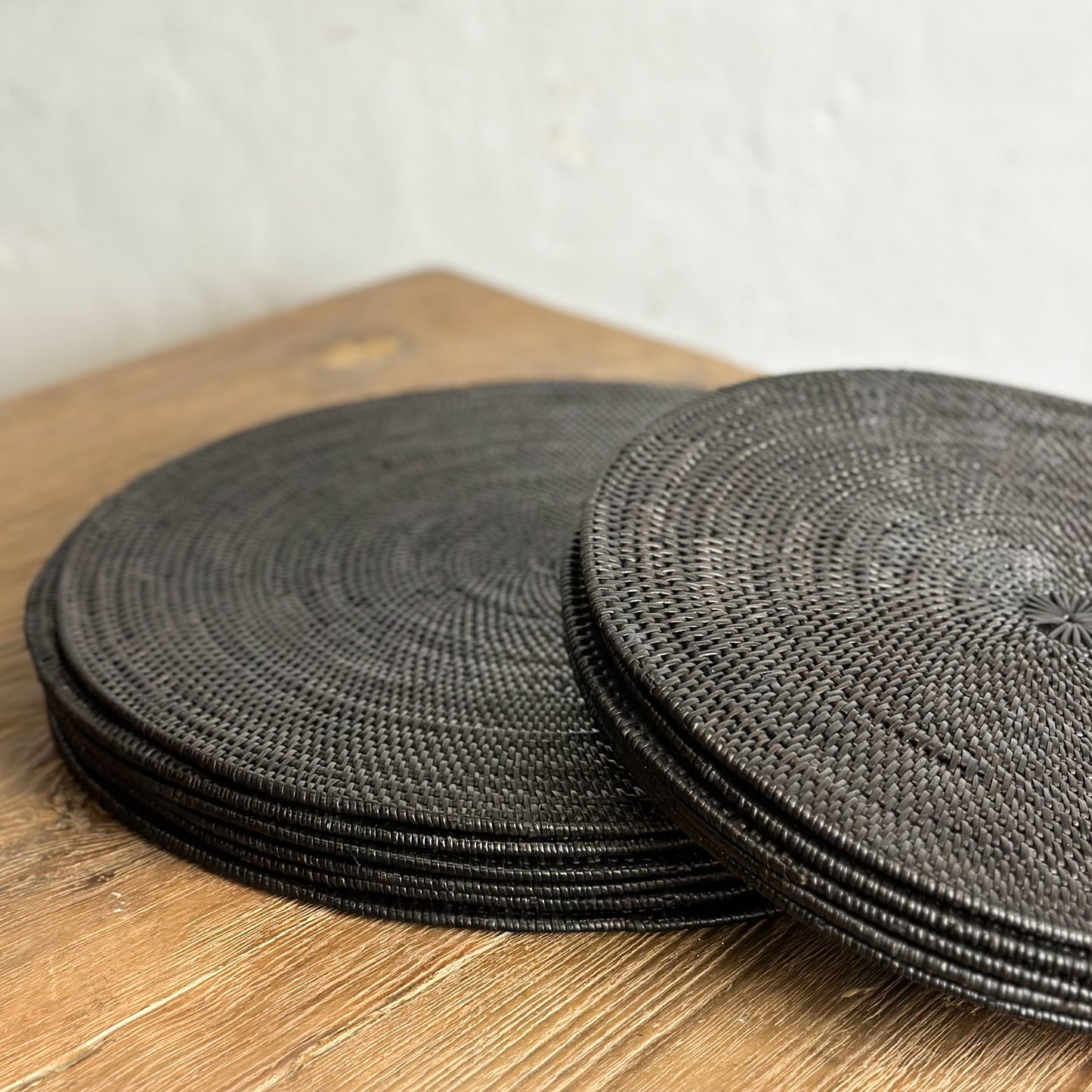 A stack of round black placemats on a wooden table surface against a white wall background.