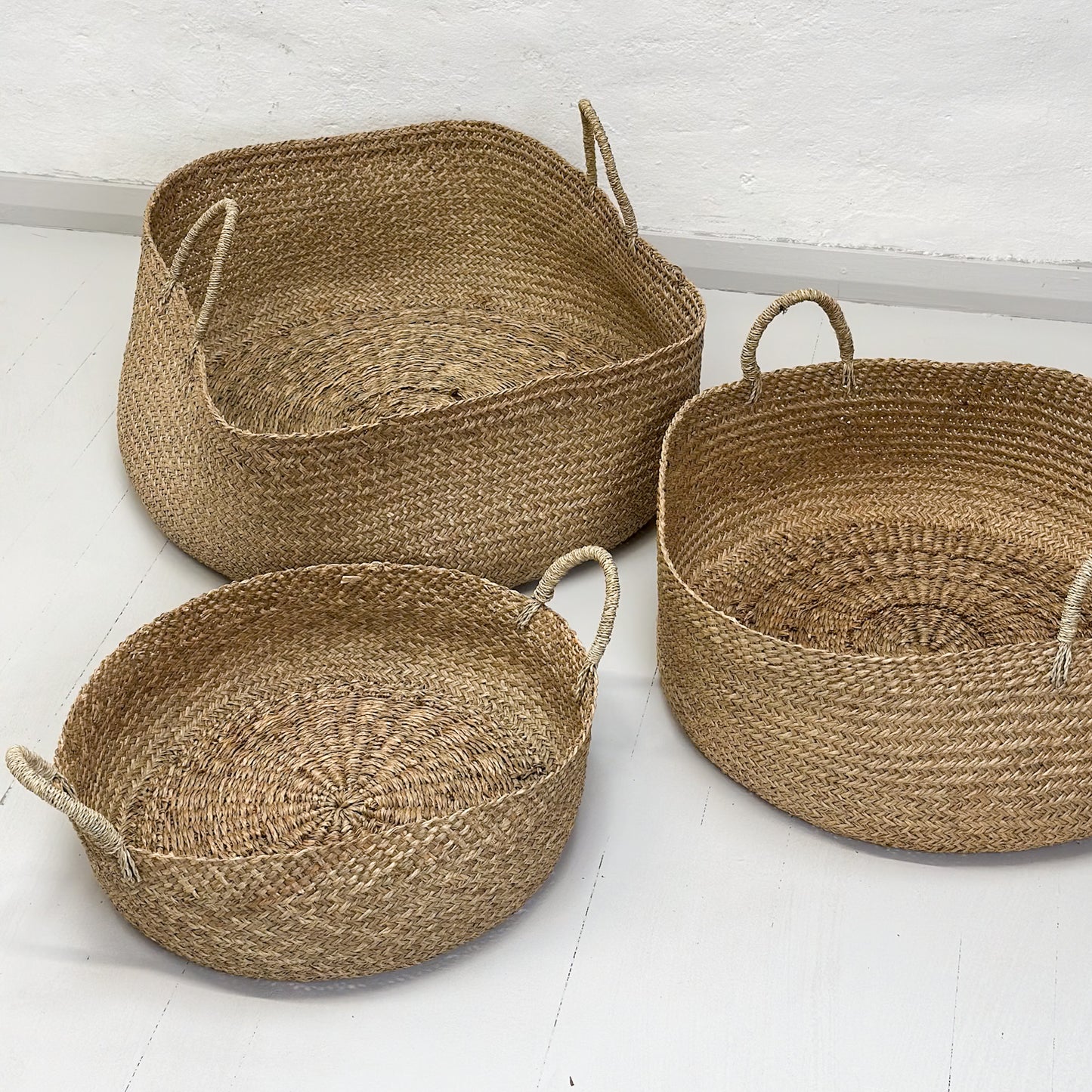 Three handcrafted wicker baskets in various sizes with natural sea grass color and texture, placed on a wooden floor against a white wall.