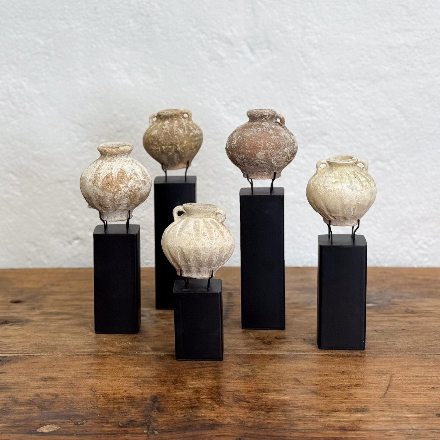 Set of five ceramic pots on black stands against a textured white wall.